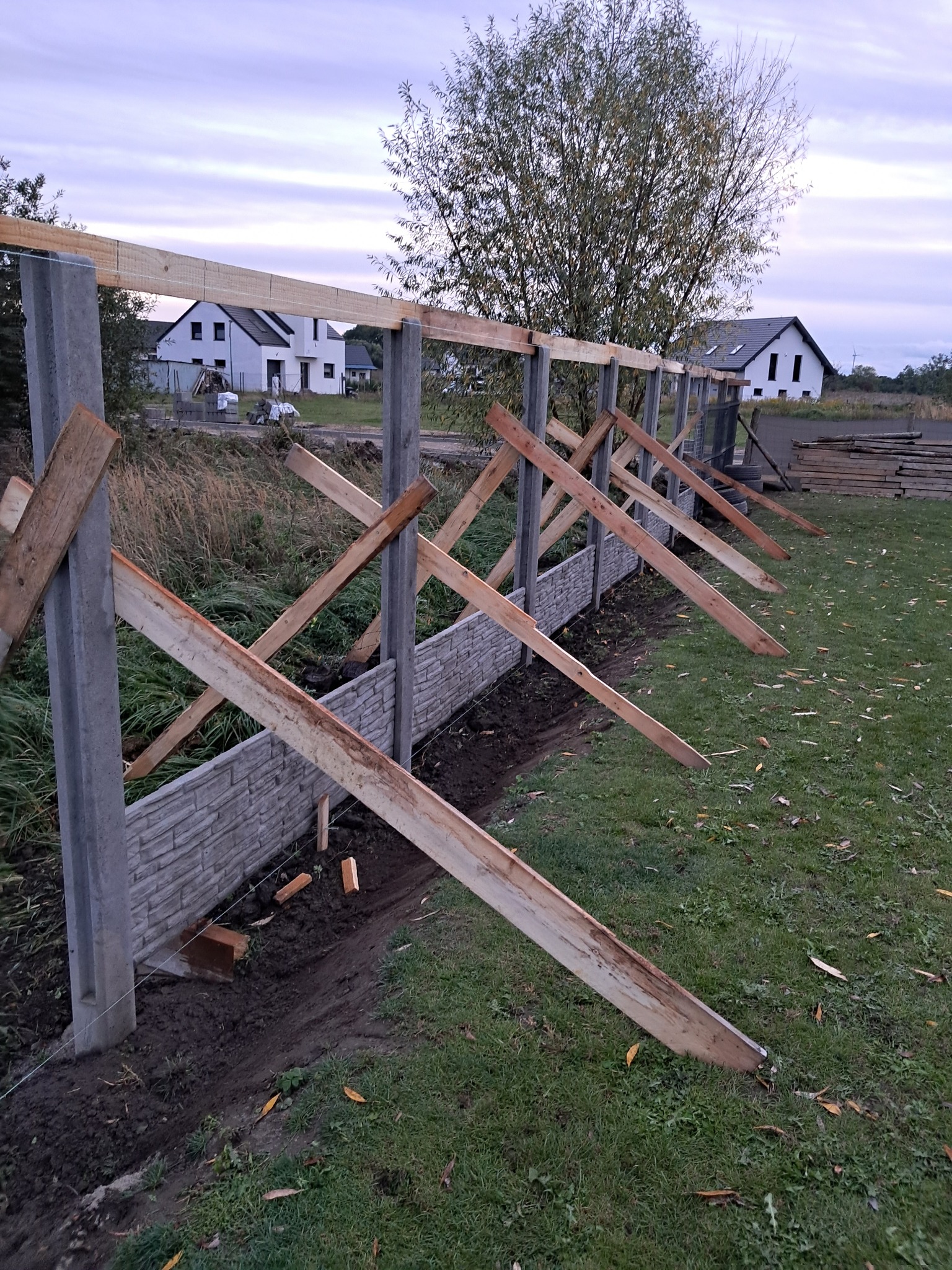 Budowa ogrodzenia z betonowych słupków i imitacji kamienia, wspartego drewnianymi belkami. W tle domy jednorodzinne i drzewo pod pochmurnym niebem.