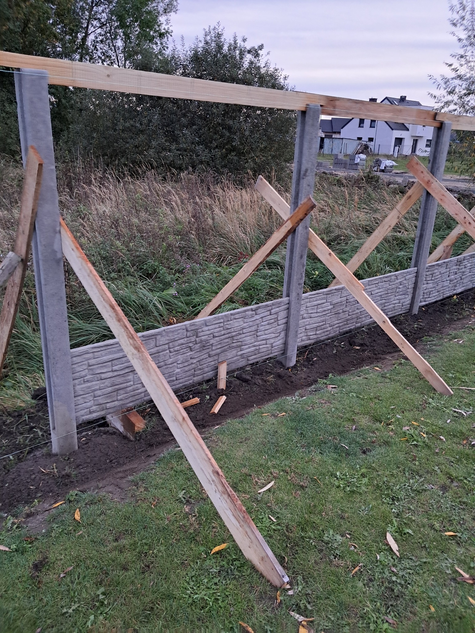 Montaż ogrodzenia panelowego z betonowych elementów imitujących cegłę. Widoczne słupki, panele i drewniane podpory. Tło stanowią trawa, drzewa i dom w oddali.