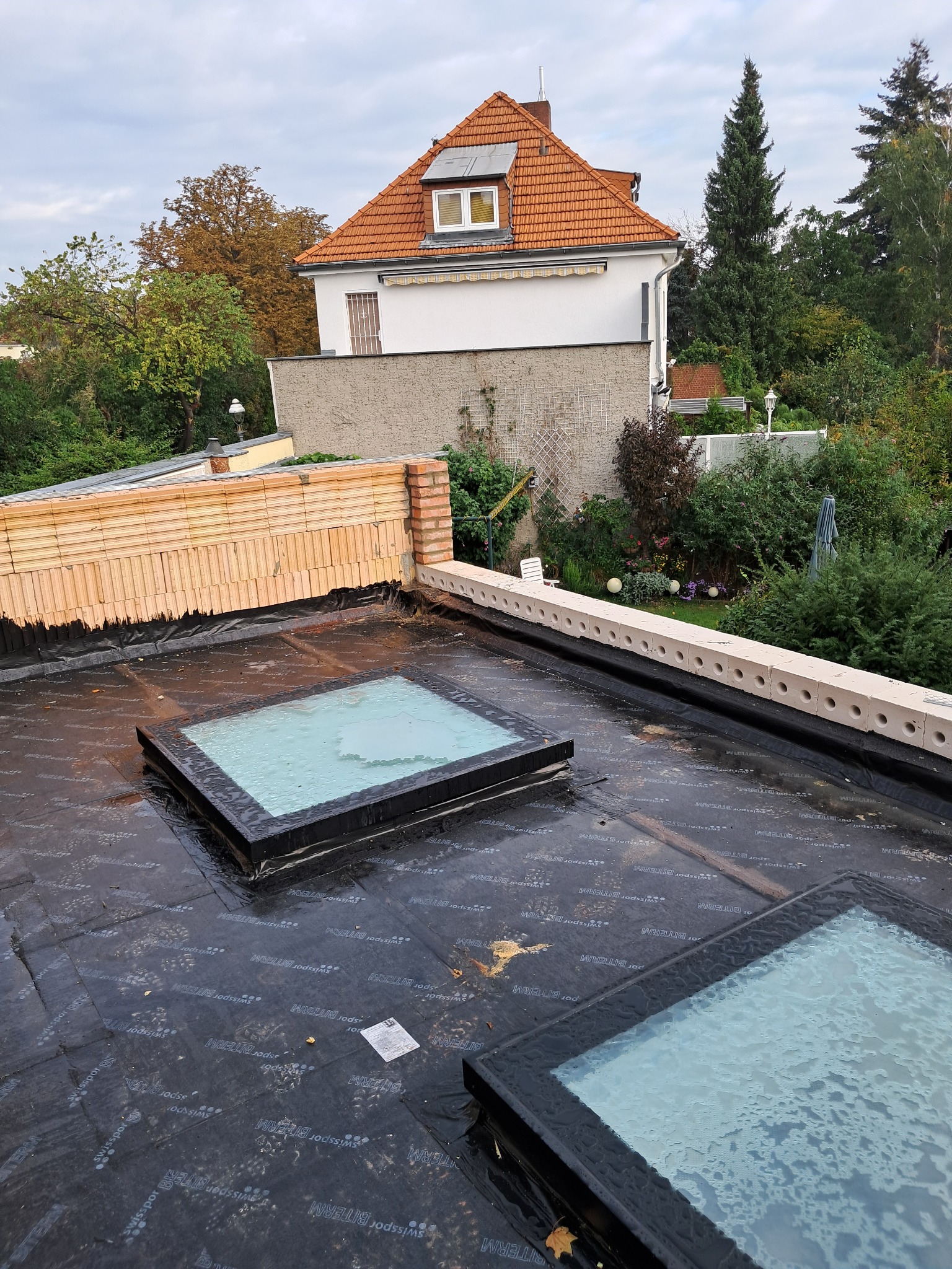 Montaż okien dachowych na płaskim dachu z papy bitumicznej. Widoczny fragment muru i budynku w tle. Dwa okna dachowe z widocznymi kroplami wody.