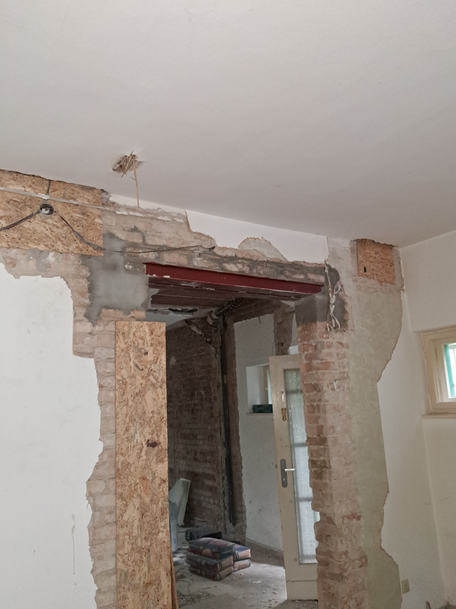 Wnętrze w trakcie remontu: odsłonięta cegła, metalowa belka nośna, płyty OSB, worki z zaprawą. Widoczne ślady po instalacjach i niedokończone ściany.