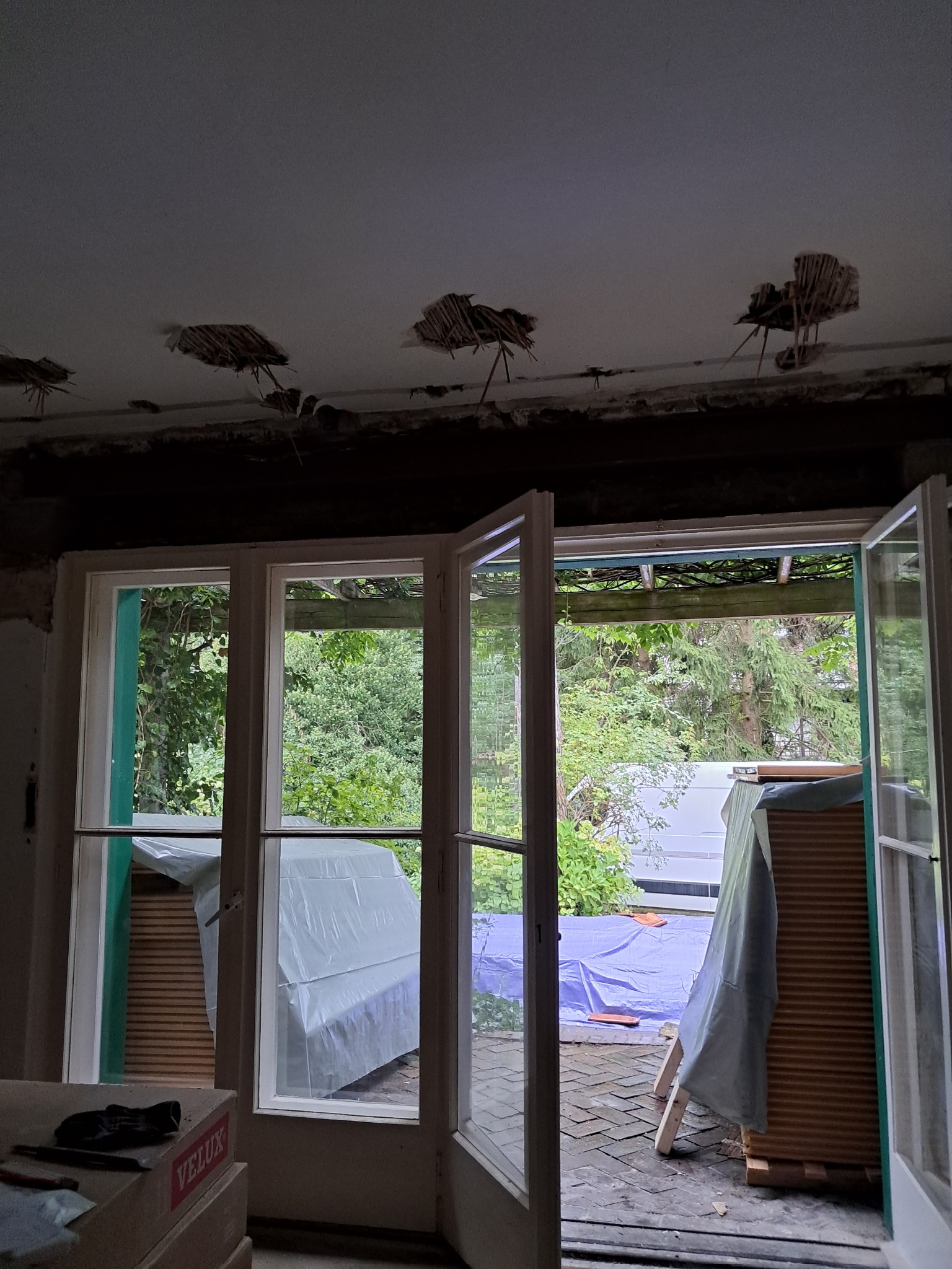 Wnętrze w trakcie remontu: otwarte okno na zewnątrz, widoczne uszkodzenia sufitu z wystającymi elementami, materiały budowlane pod folią ochronną.