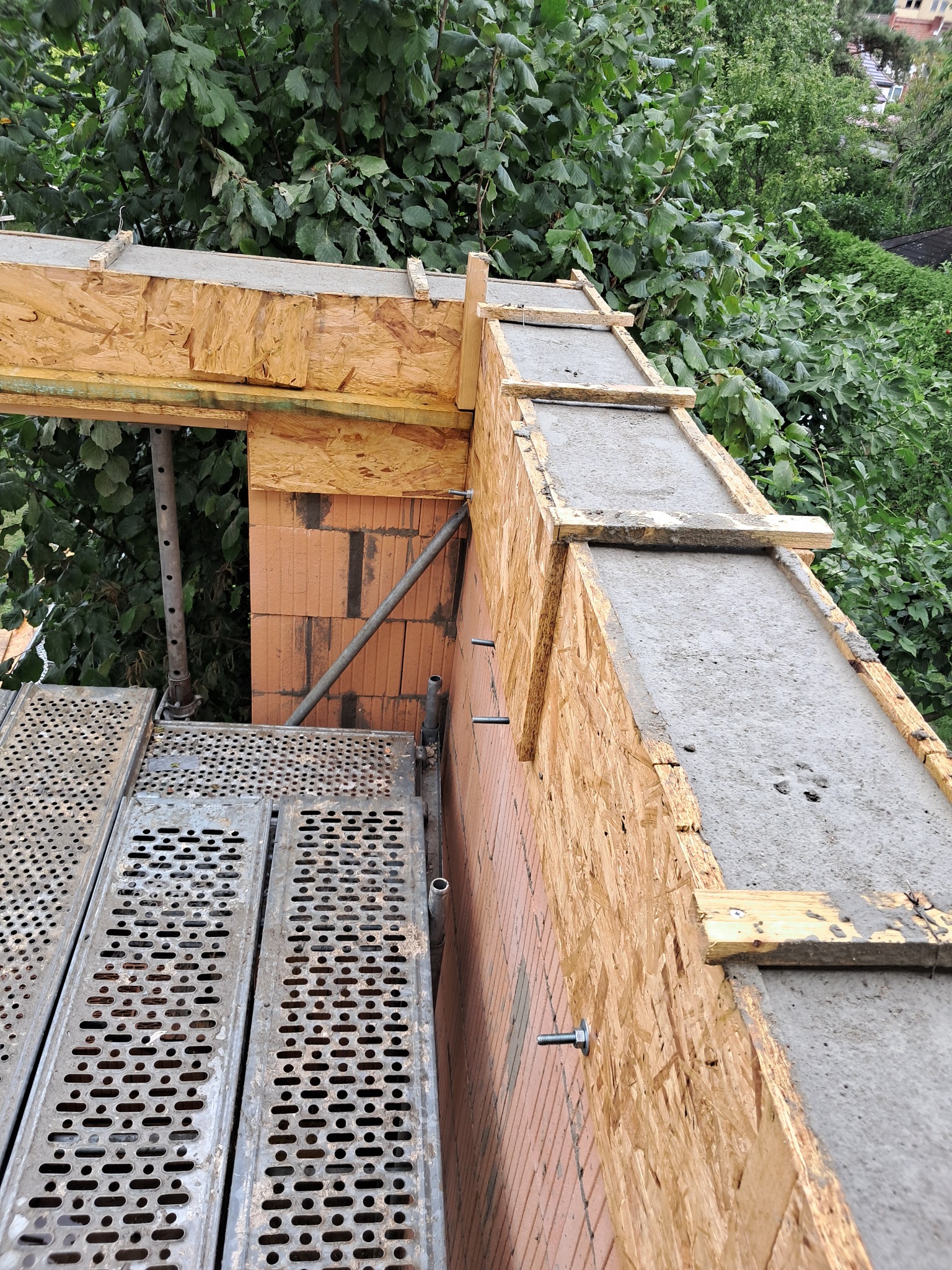 Wysoki kąt: mur z cegły, szalunek z płyty OSB wypełniony betonem, rusztowanie i zieleń w tle. Widoczne śruby mocujące szalunek. Etap budowy.