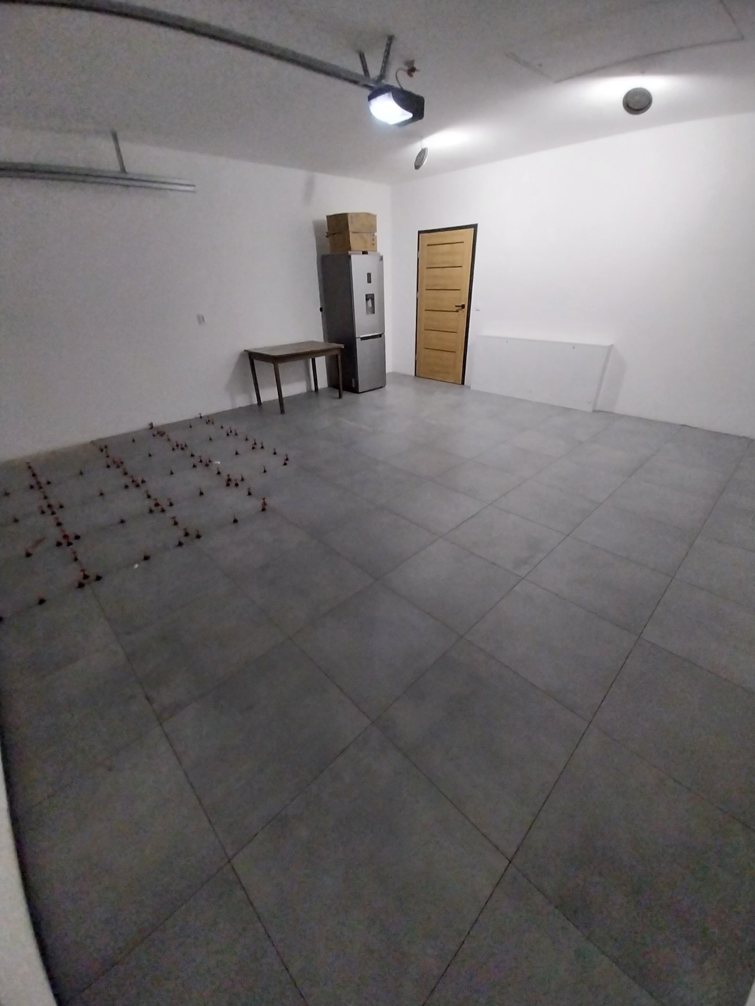 Szare płytki podłogowe w jasnym garażu, częściowo ułożone, z czerwonymi krzyżykami dystansowymi. Widoczny stół, lodówka i drzwi w tle. Oświetlenie sufitowe.