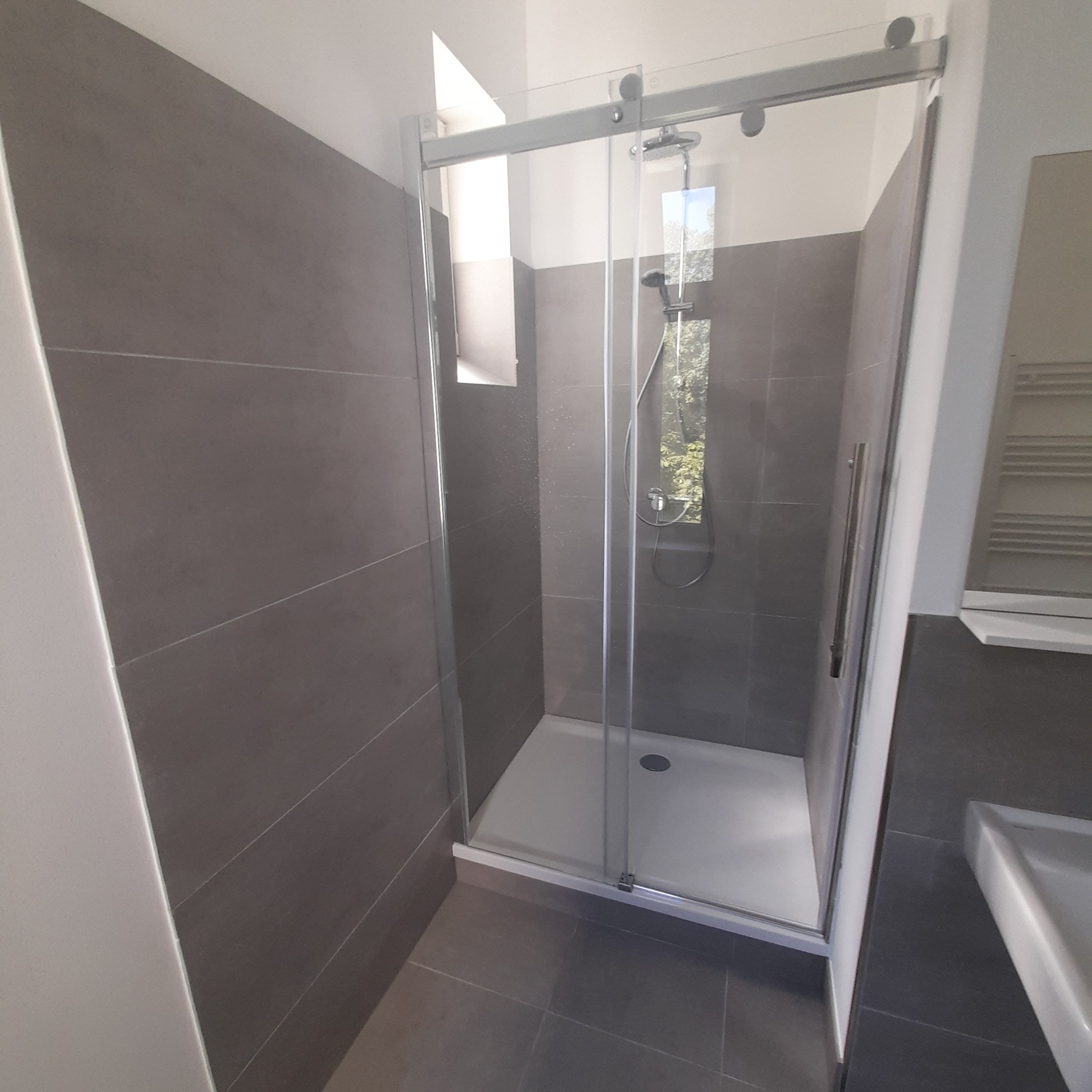 Nowoczesna kabina prysznicowa z przesuwanymi drzwiami, szare płytki na ścianach i podłodze. Widok na zewnątrz przez szybę prysznica.
