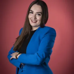 Uśmiechnięta brunetka w niebieskim garniturze, z zegarkiem na ręce, pozuje na tle koralowego studia. Profesjonalny wizerunek.