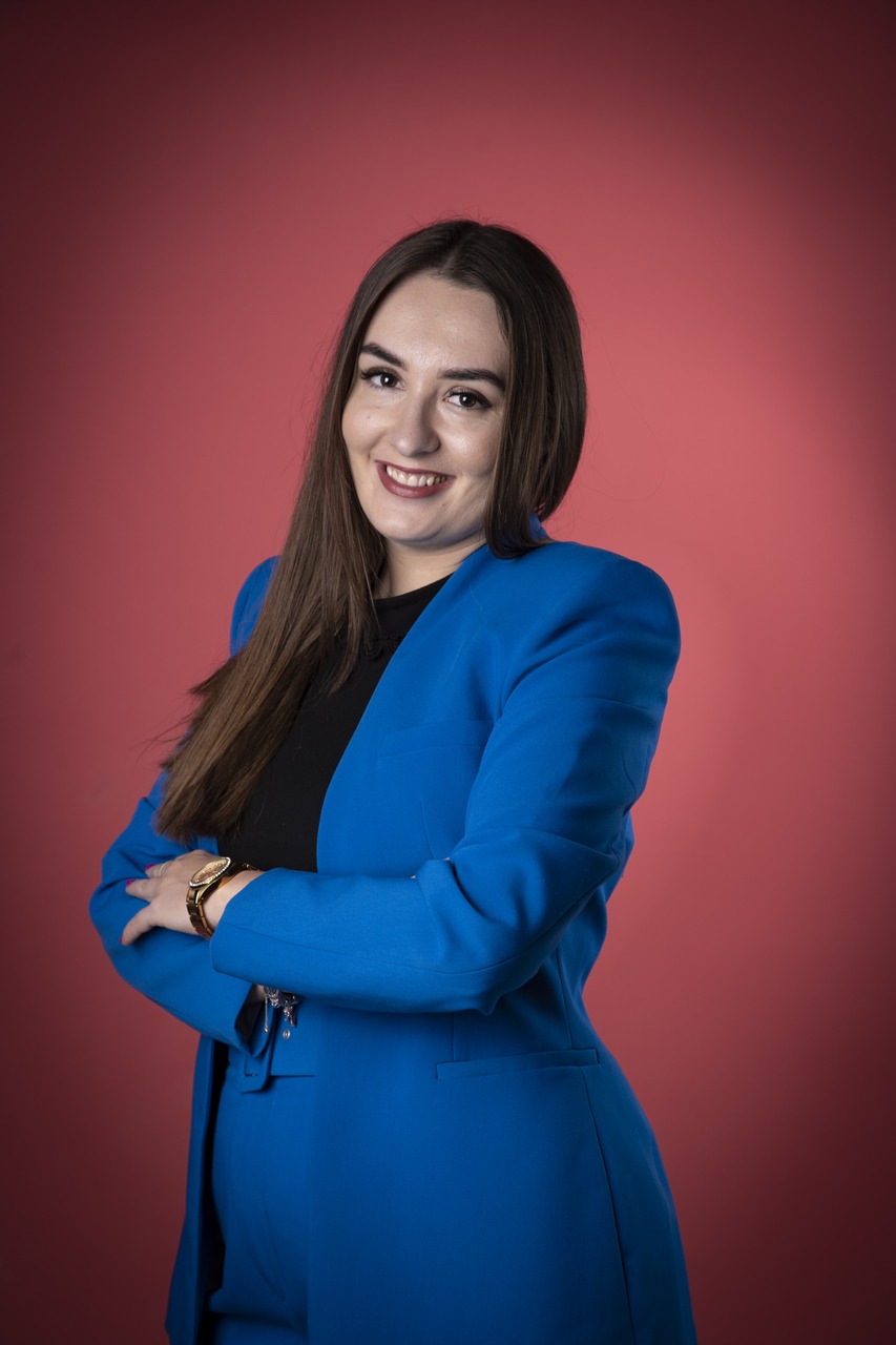 Uśmiechnięta brunetka w niebieskim garniturze, z zegarkiem na ręce, pozuje na tle koralowego studia. Profesjonalny wizerunek.