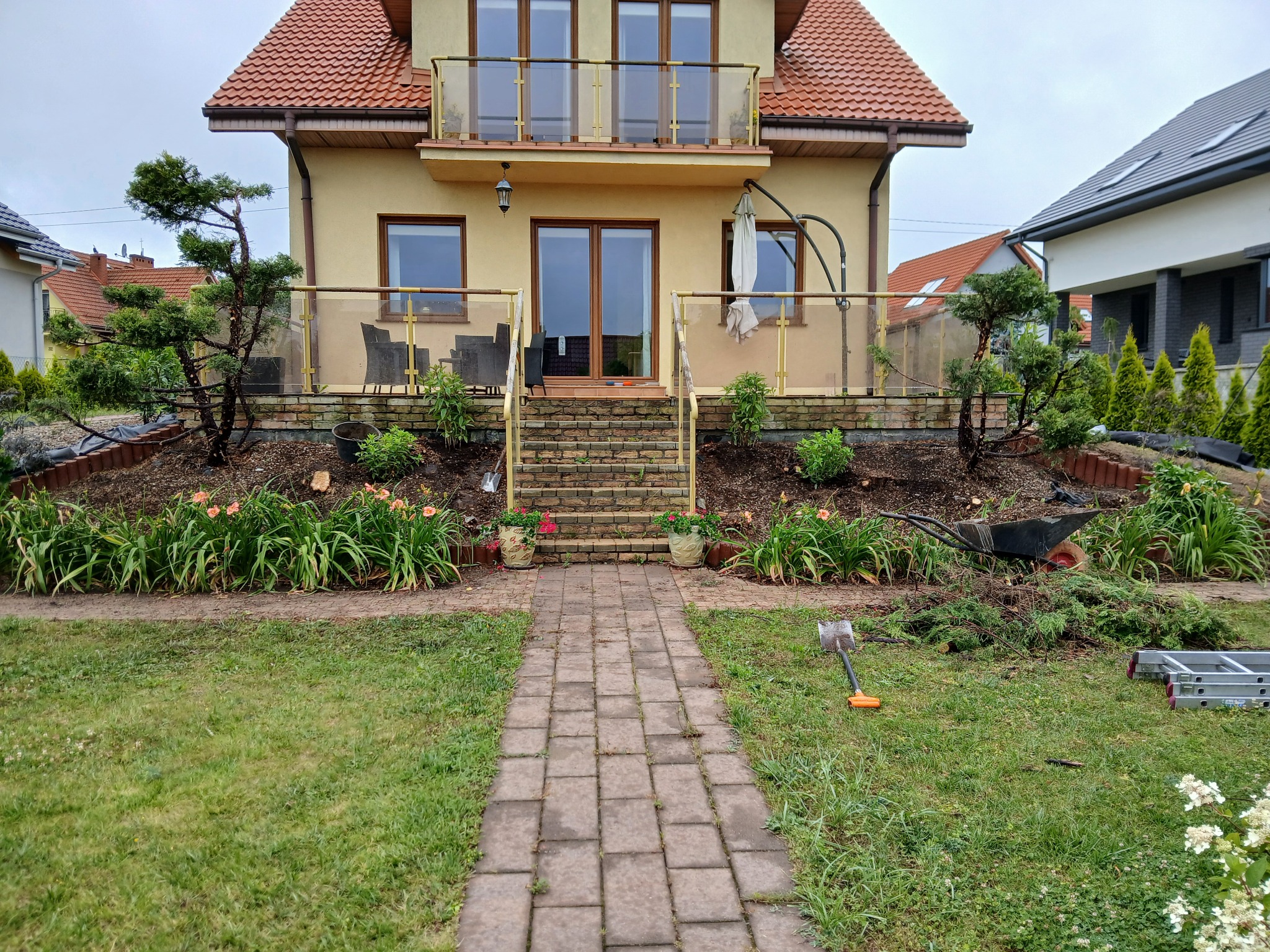Ogród po przycięciu drzew i krzewów przed domem. Widoczne narzędzia ogrodnicze, taczka i drabina. Ułożona kostka prowadzi do wejścia.