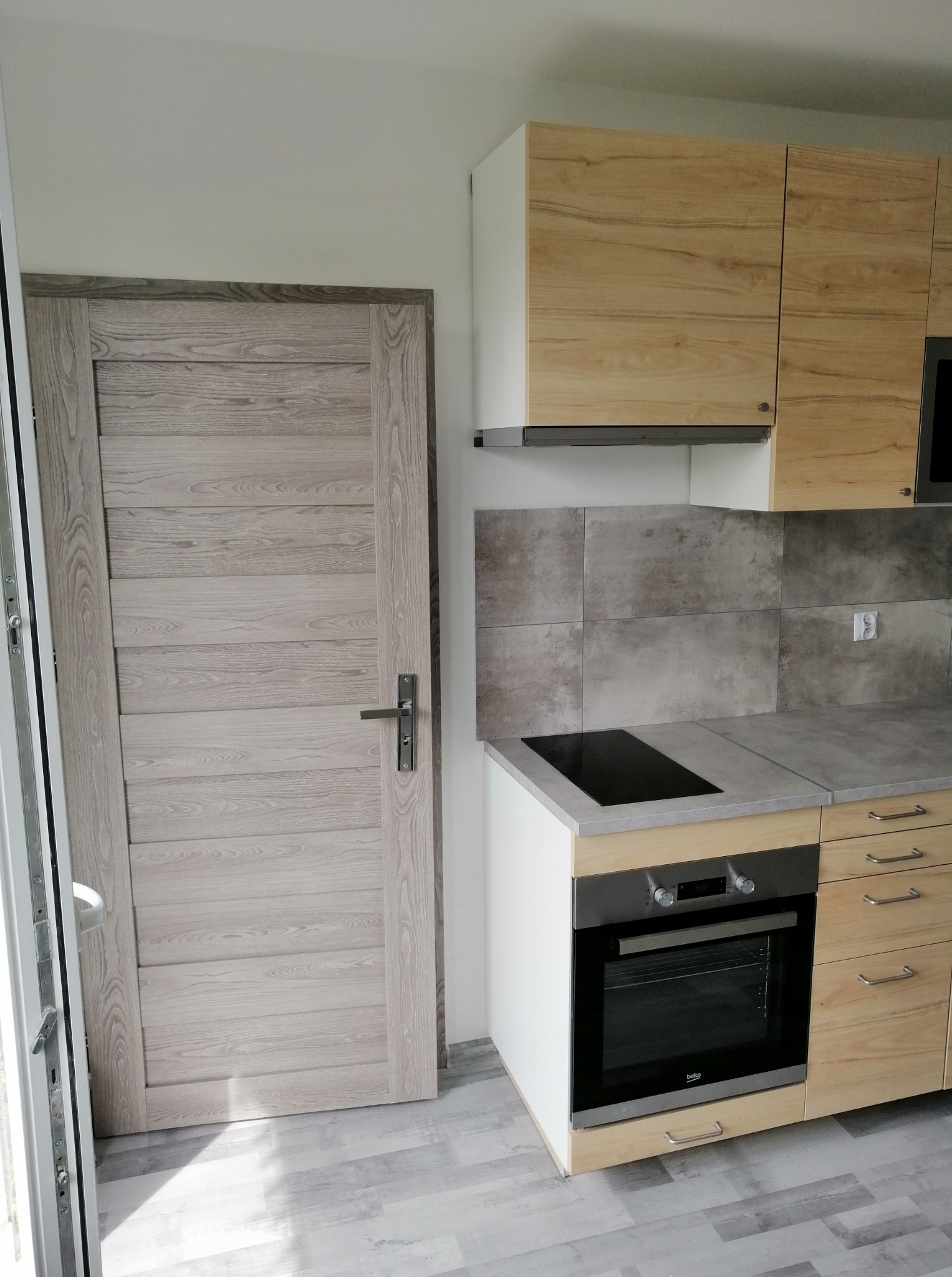 Jasna kuchnia po remoncie z drewnopodobnymi frontami szafek, szarą płytą indukcyjną i drzwiami w odcieniach szarości, widok z poziomu podłogi.