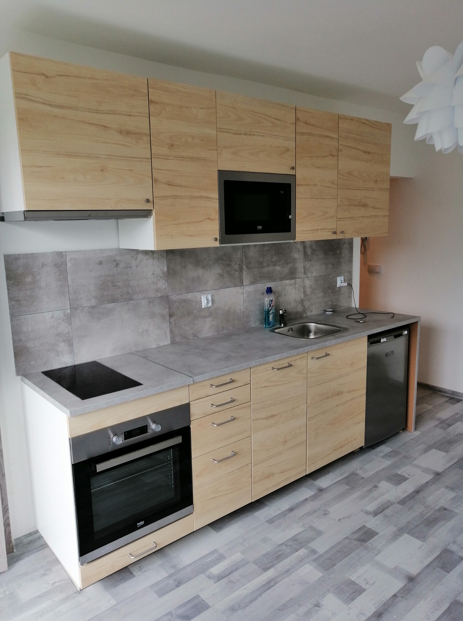 Jasna kuchnia z drewnianymi frontami, szarym blatem i płytkami. Widoczny piekarnik, płyta indukcyjna i mikrofalówka. Minimalistyczny styl, nowoczesne oświetlenie.