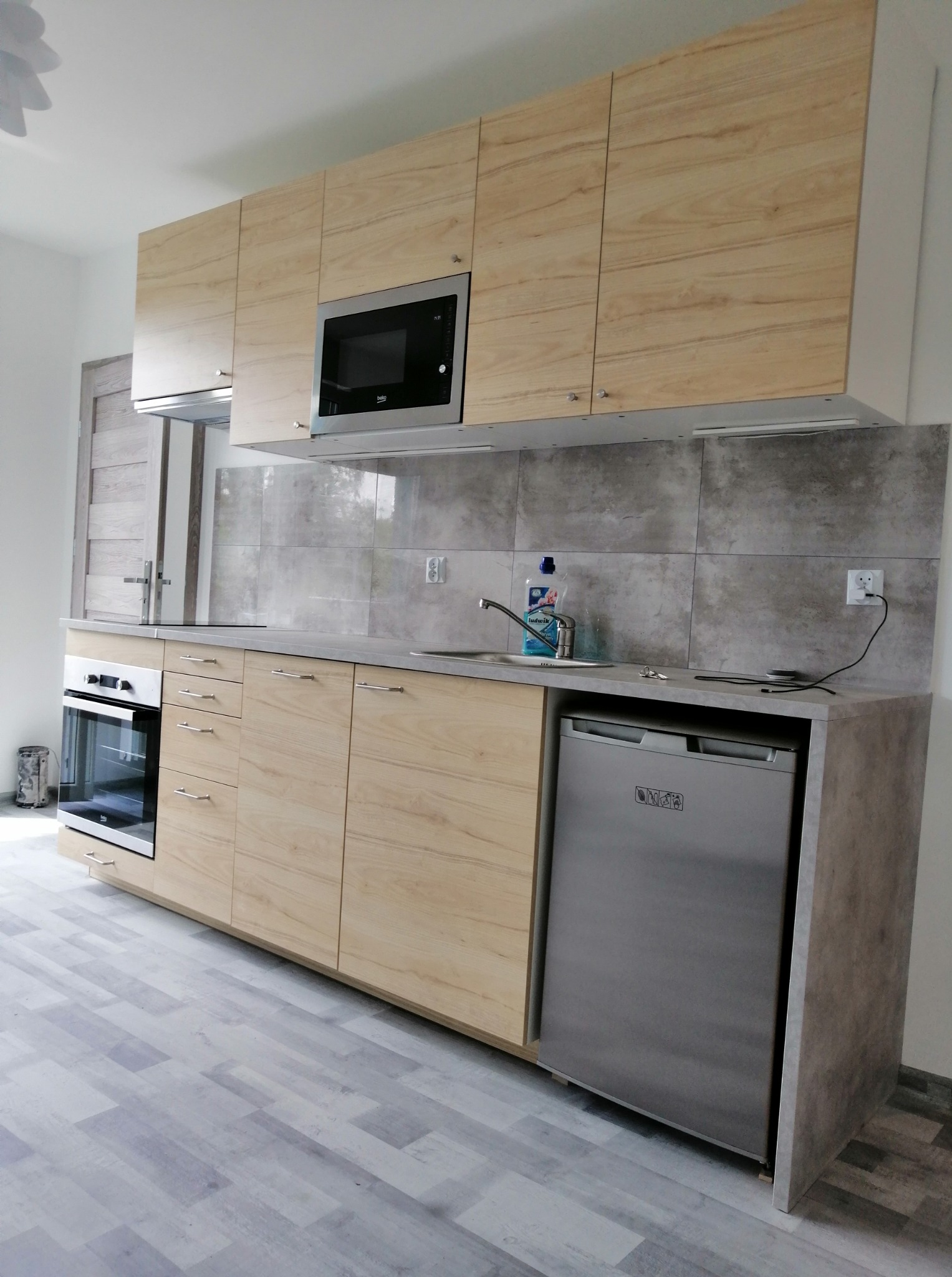 Jasna, nowoczesna kuchnia z drewnianymi frontami, srebrnym sprzętem AGD i szarą glazurą. Widoczny zlew, płyta, piekarnik i lodówka. Podłoga w odcieniach szarości.