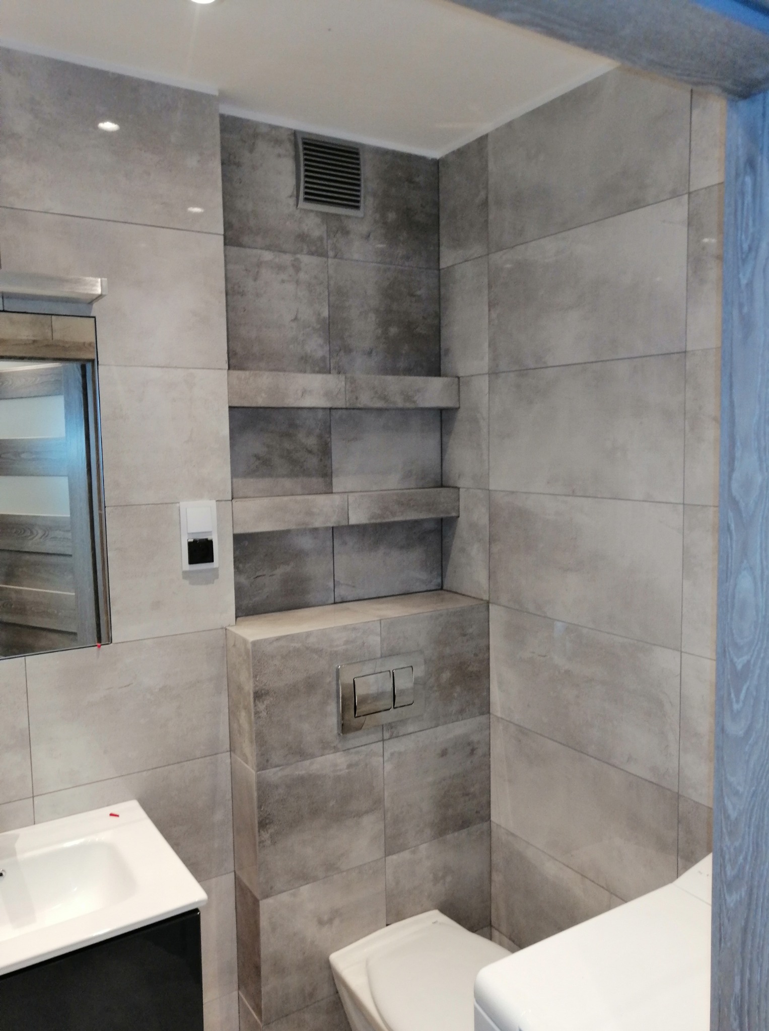 Wykończona łazienka w szarościach: płytki imitujące kamień, zabudowa WC z przyciskiem, umywalka z szafką i lustro. Półki w ścianie, wentylacja.