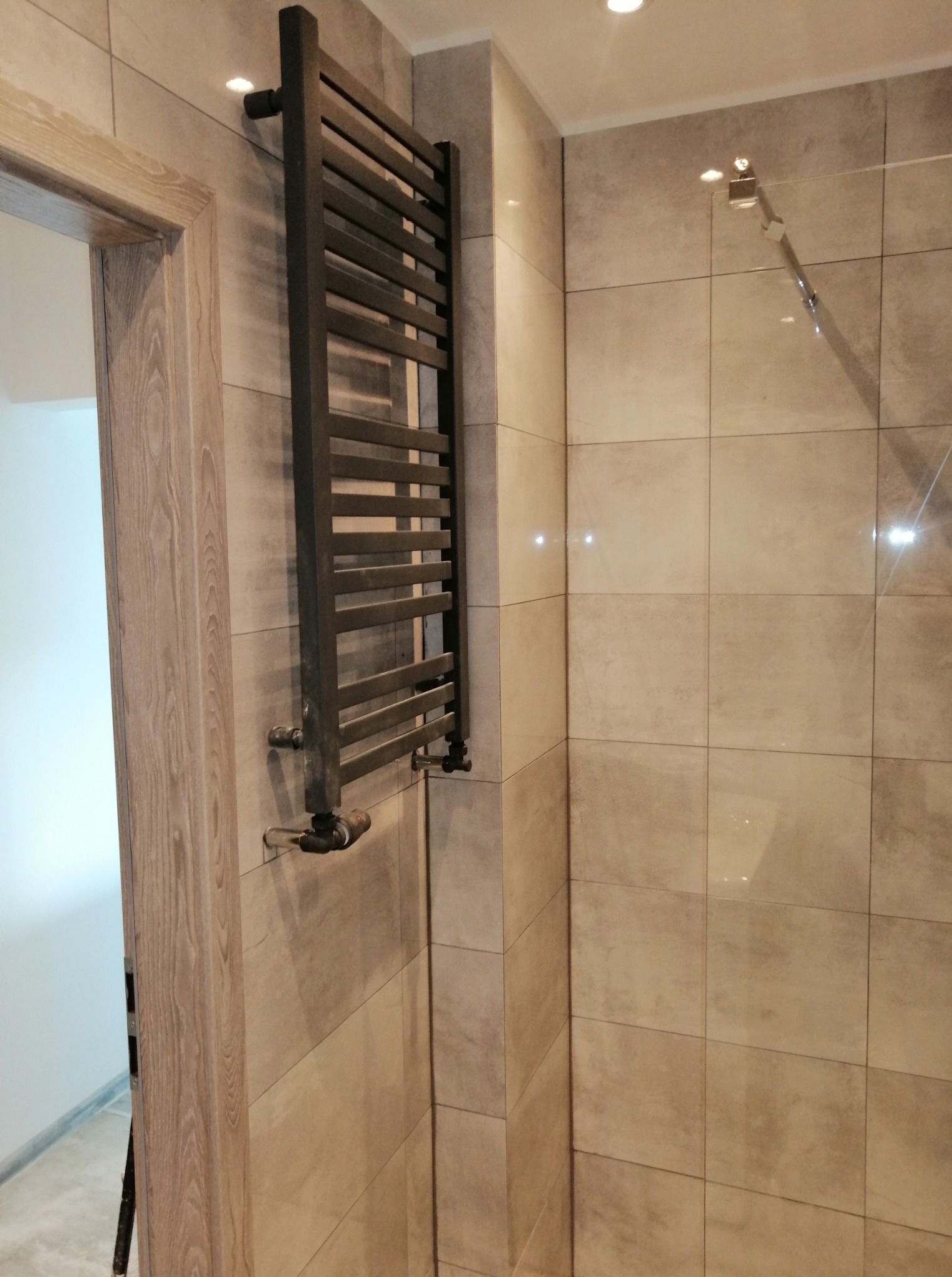 Nowoczesna łazienka z grafitowym grzejnikiem drabinkowym na beżowych płytkach. Widoczny fragment kabiny prysznicowej i drzwi.