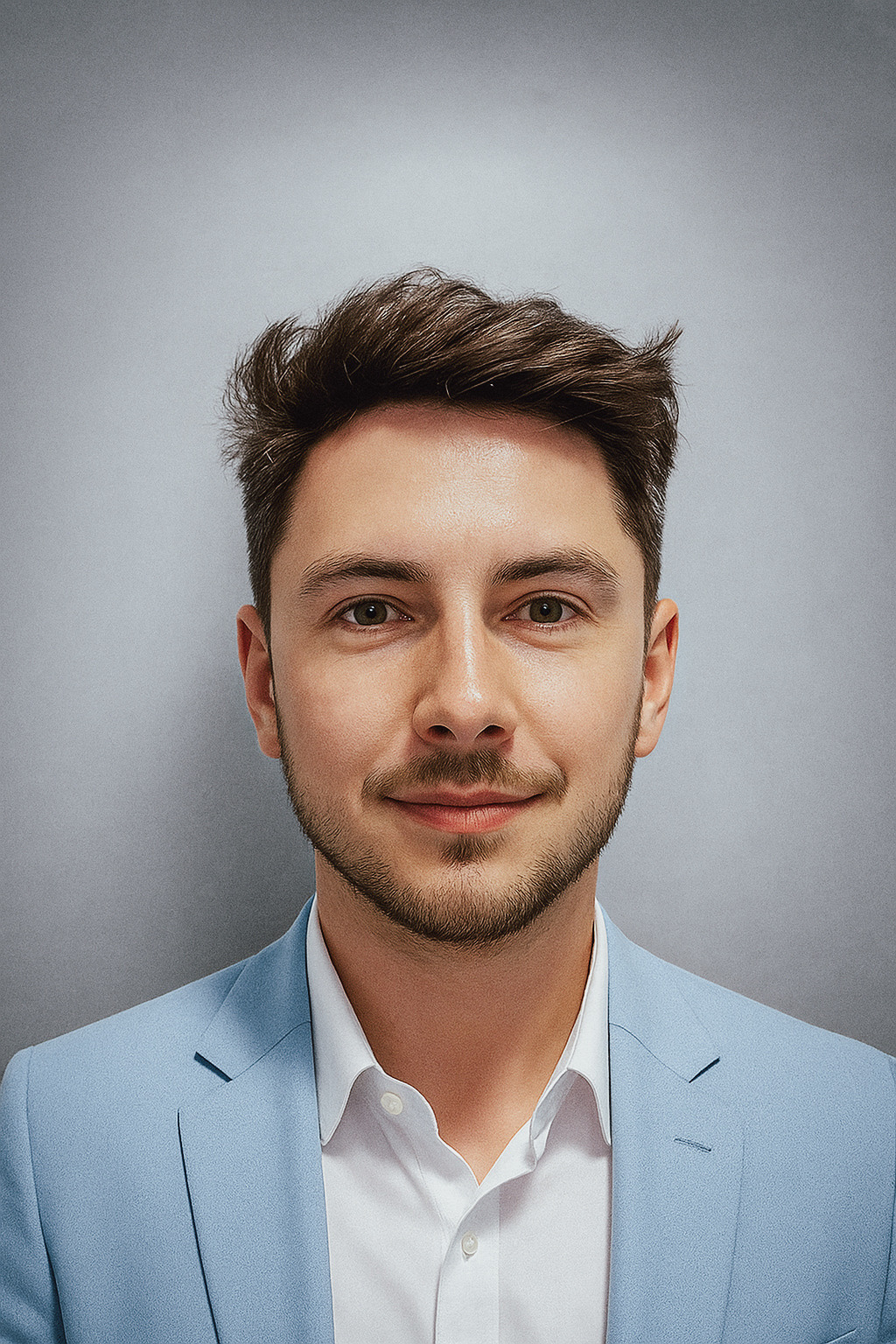 Mężczyzna w błękitnej marynarce i białej koszuli, uśmiechnięty, na szarym tle. Portret biznesowy z lekkim zarostem i brązowymi włosami.