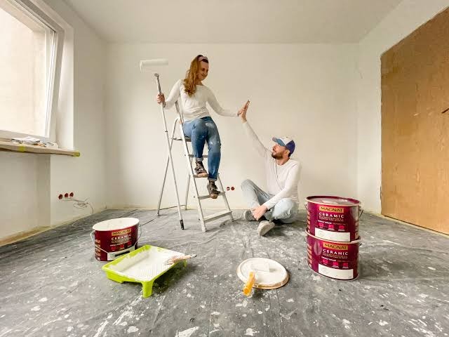 Para daje sobie 'piątkę' w trakcie malowania ścian w pomieszczeniu. Kobieta na drabinie z wałkiem. Widoczne puszki z farbą i akcesoria malarskie.