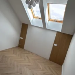 Jasne, nowe panele ułożone w jodełkę w pokoju na poddaszu z oknami dachowymi i nowoczesną lampą. Widoczne drewniane panele na ścianach z gniazdkami.