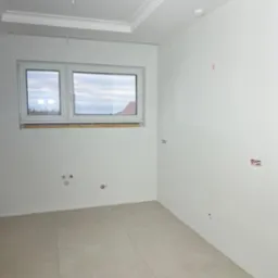 Wnętrze po remoncie: gładkie, białe ściany, jasne płytki podłogowe, okno z widokiem na zewnątrz. Widoczne punkty instalacji elektrycznej i wodnej.