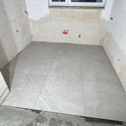 InHome AM - Ułożone jasne płytki podłogowe w pokoju z oknem, ściany przygotowane do malowania, widoczny odkurzacz. Prace wykończeniowe w trakcie realizacji.