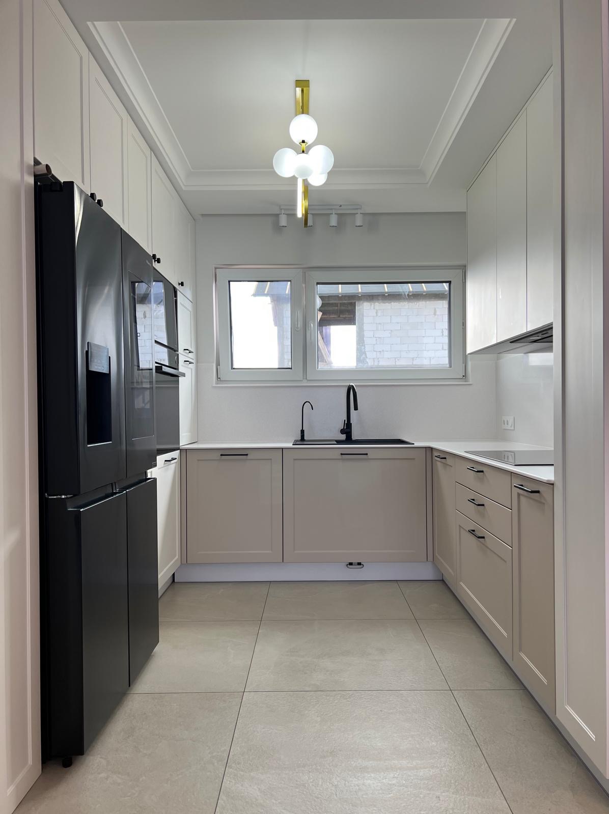 Nowoczesna kuchnia w Elblągu z beżowymi szafkami i czarnymi akcentami: bateria, zlew, lodówka. Białe szafki górne i płytki podłogowe. Lampa sufitowa w stylu glamour.