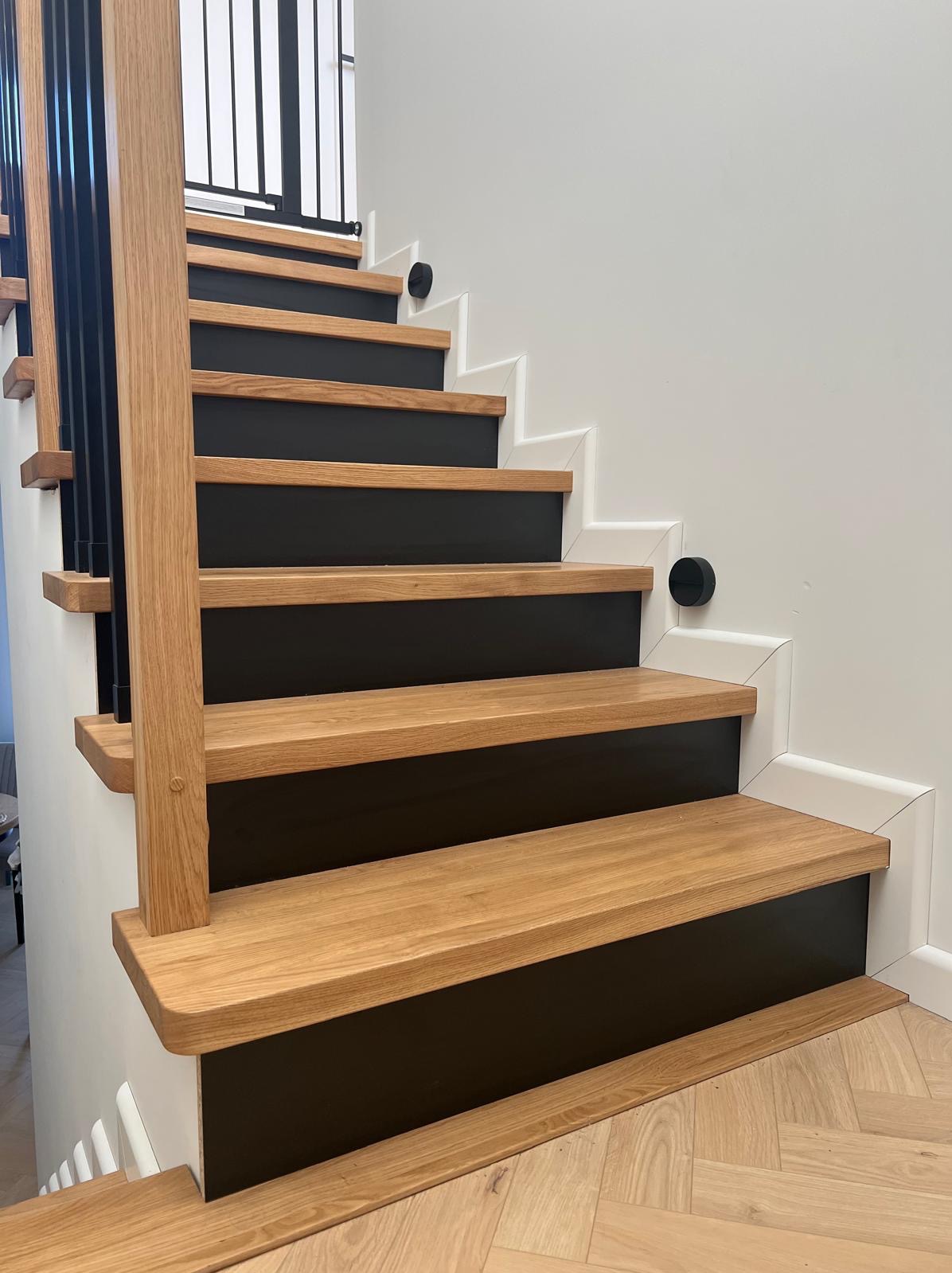 Drewniane schody z czarnymi podstopnicami i białymi listwami przypodłogowymi, zabezpieczone bramką na górze. Parkiet ułożony w jodełkę na podłodze.