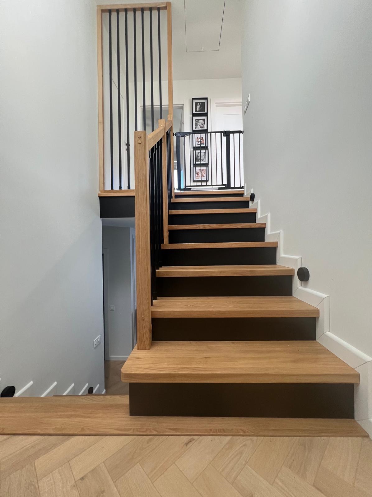 Drewniane schody z czarnymi podstopnicami i balustradą, widok z dołu. Parkiet ułożony w jodełkę. Na ścianie zdjęcia w ramkach.