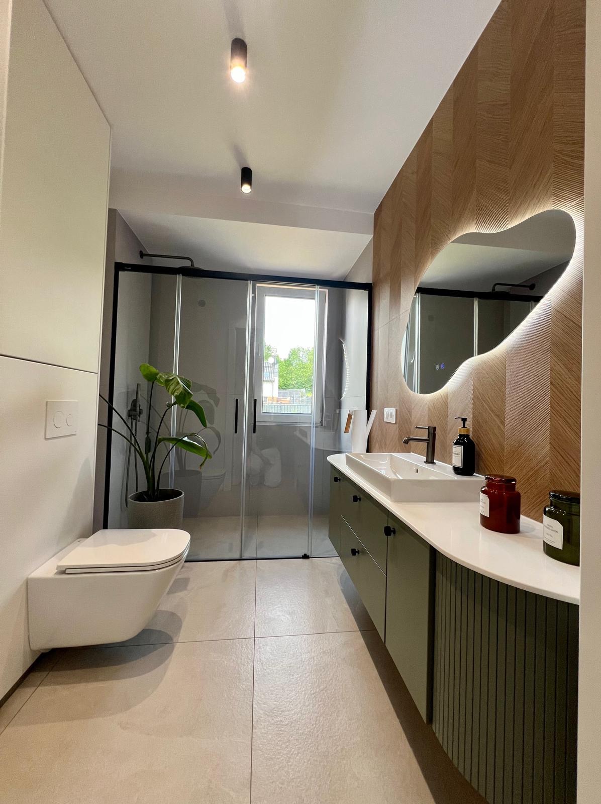 Nowoczesna łazienka z kabiną prysznicową, umywalką na szafce w kolorze oliwkowym i dekoracyjnym lustrem z podświetleniem LED na ścianie z drewnianą okładziną.