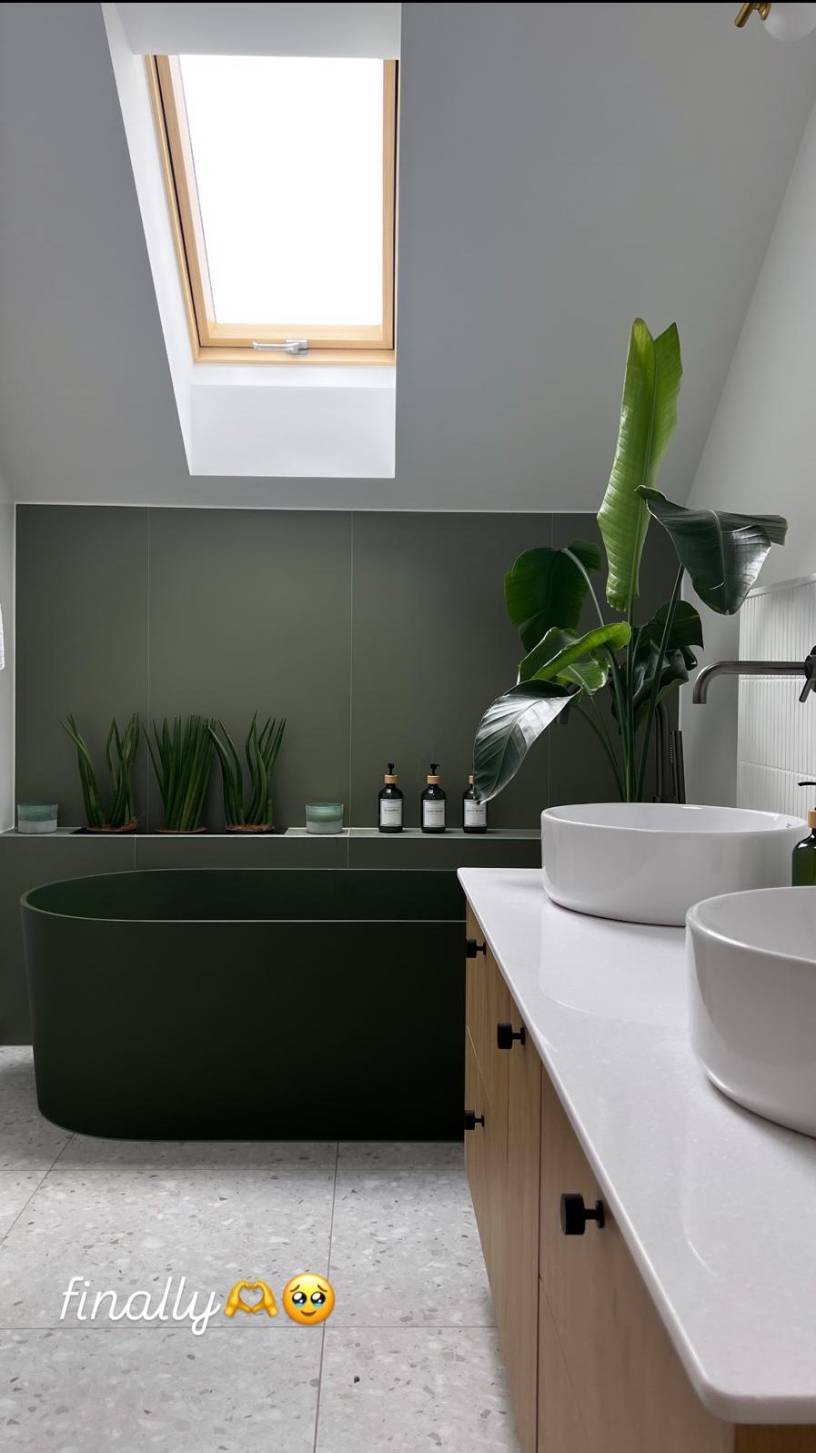 Stylowa łazienka w odcieniach zieleni z wolnostojącą wanną, roślinami i oknem dachowym, wykończona w nowoczesnym stylu z elementami drewnianymi.