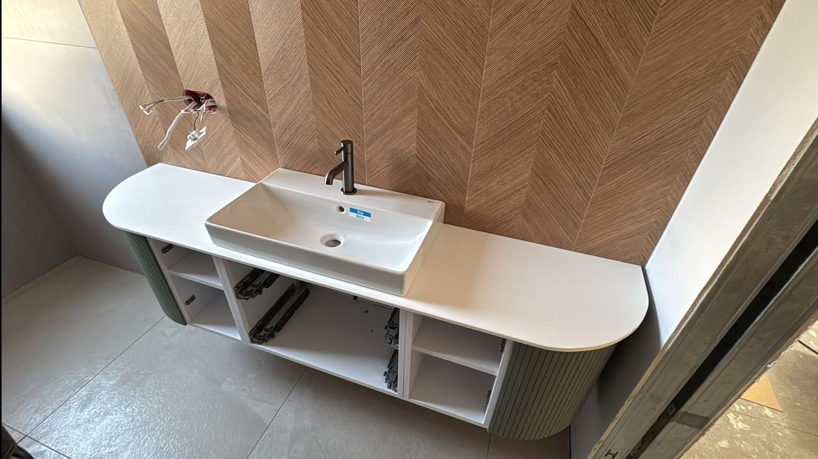 Nowoczesna łazienka z umywalką nablatową i szafką w kolorze zieleni. Na ścianie dekoracyjne płytki w jodełkę. Widoczne niedokończone podłączenie elektryczne.