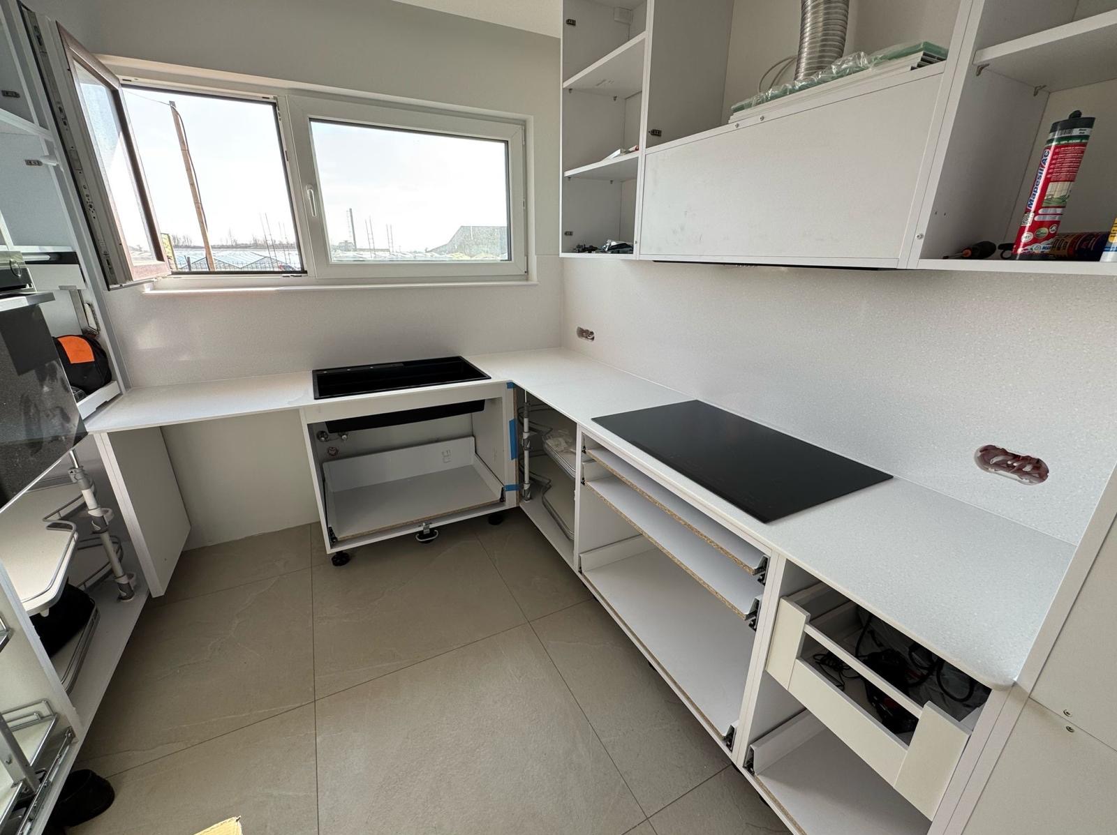 Biała, częściowo zamontowana kuchnia z widokiem na port przez okno. Fragment blatu z płytą indukcyjną. Etap montażu, widoczne narzędzia i akcesoria.