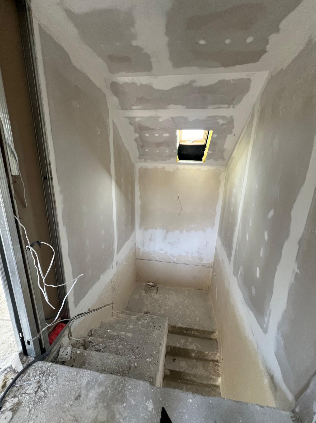 Klatka schodowa w trakcie szpachlowania. Widoczne ściany z nałożoną masą, sufit z oknem dachowym i fragment metalowej konstrukcji. Etap przygotowania pod malowanie.