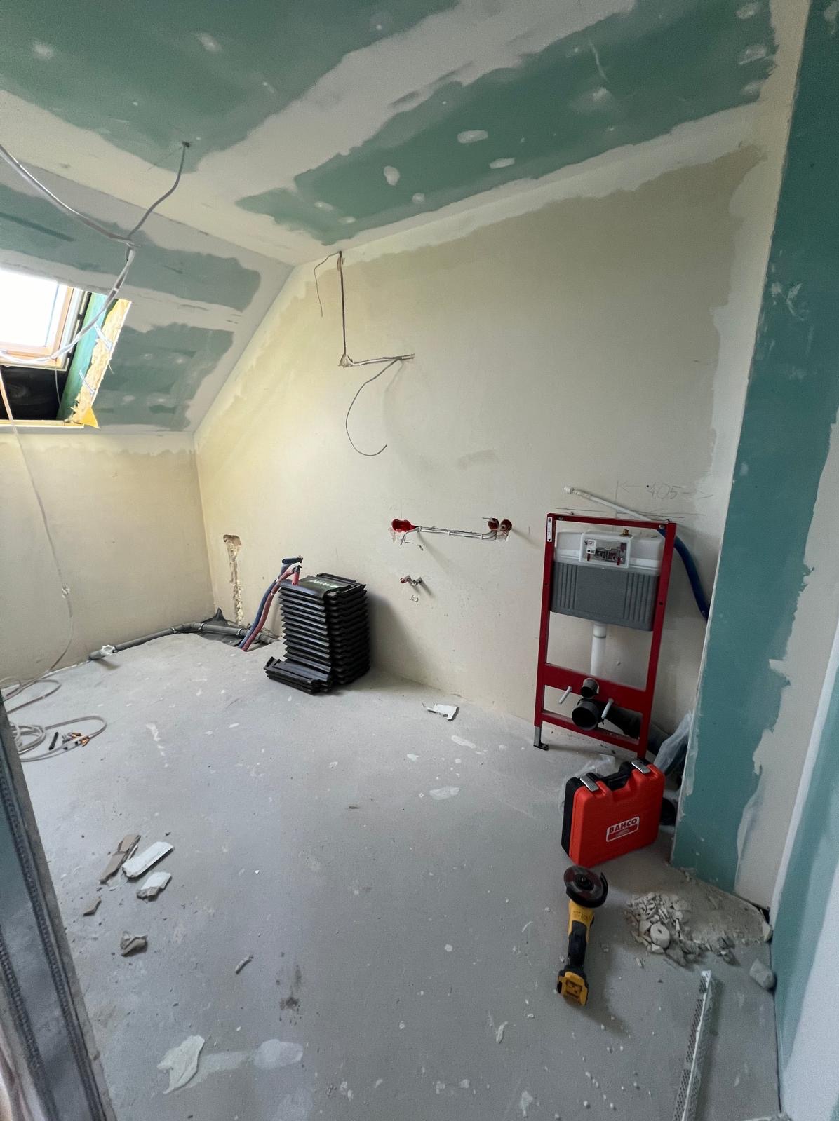 Pokój na poddaszu w trakcie prac wykończeniowych: ściany przygotowane pod malowanie, widoczna instalacja sanitarna, narzędzia i materiały budowlane.