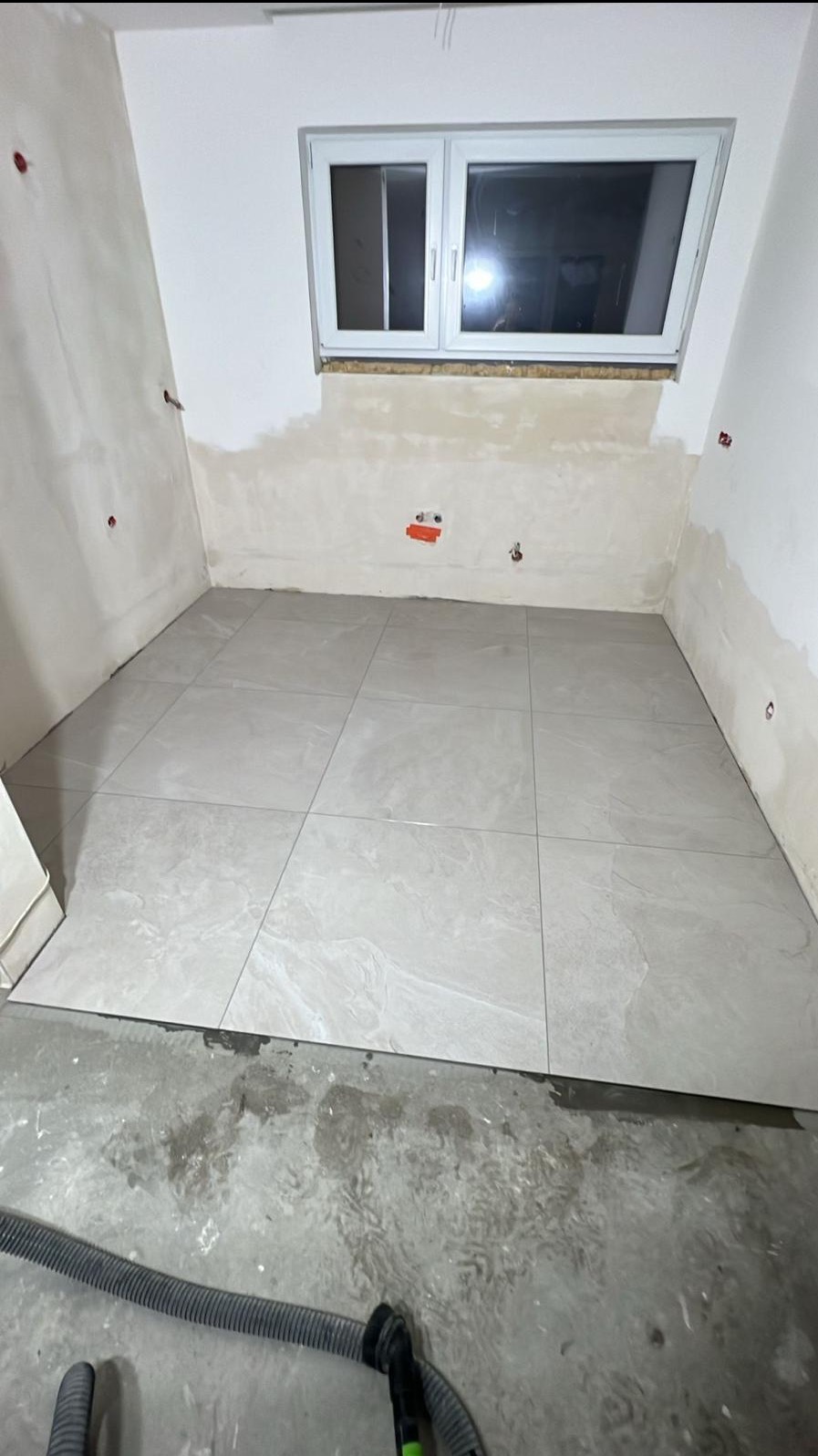 Ułożone jasne płytki podłogowe w pokoju z oknem, ściany przygotowane do malowania, widoczny odkurzacz. Prace wykończeniowe w trakcie realizacji.