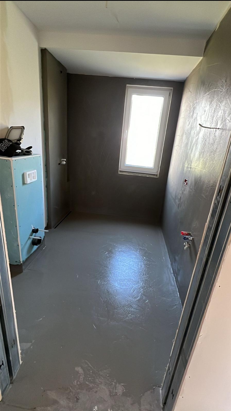 Szara łazienka w trakcie realizacji, widoczne surowe ściany, wylewka na podłodze, okno i instalacja sanitarna. W tle narzędzia.