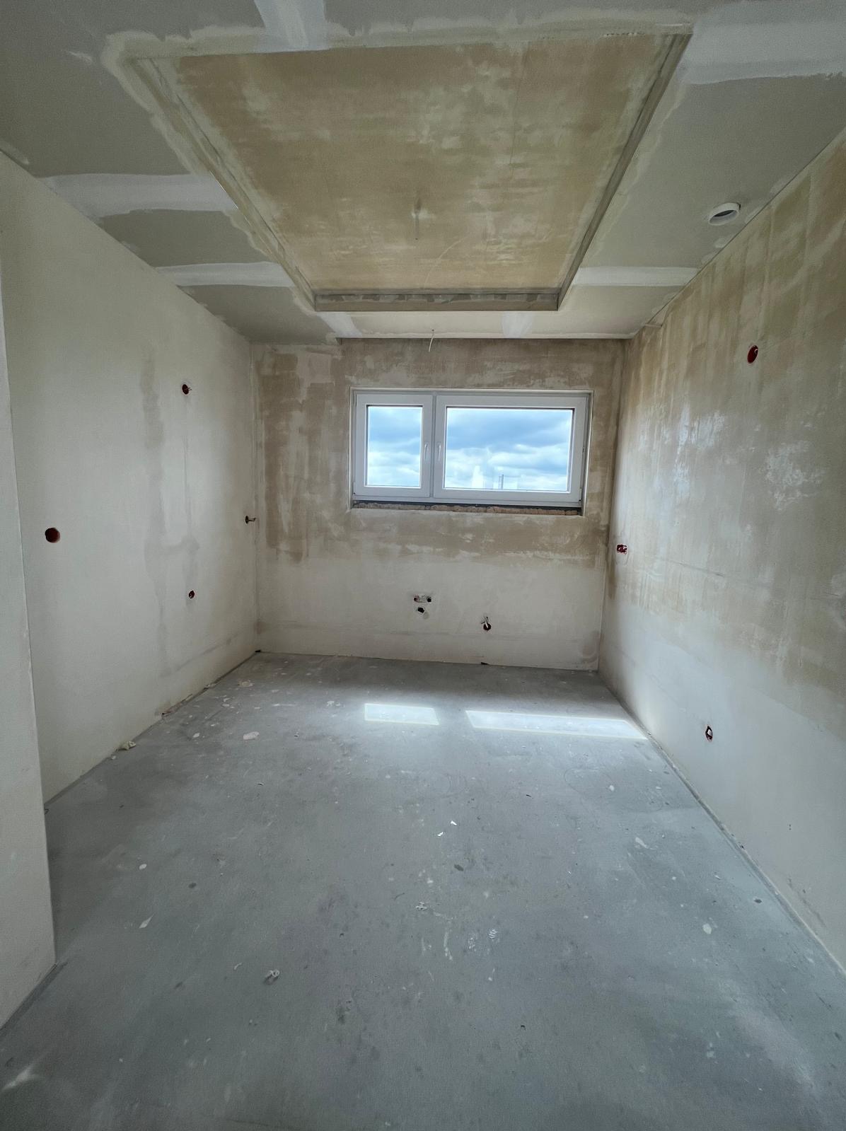 Pokój w trakcie prac wykończeniowych: surowe ściany z widocznymi punktami elektrycznymi, betonowa podłoga i podwieszany sufit z elementami szpachlowania.