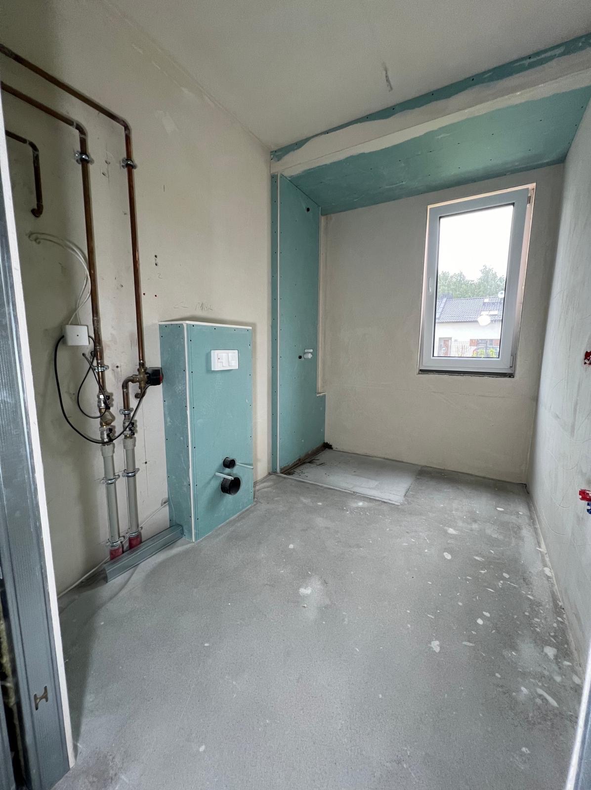 Wnętrze łazienki w trakcie prac wykończeniowych: widoczne rury, zabudowa z płyt gipsowych, okno. Szare ściany i podłoga w surowym stanie.