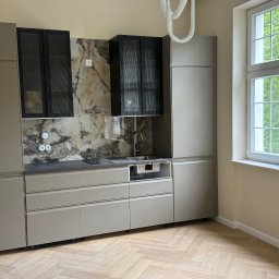 Bartosz Górski - Ukończona kuchnia w odcieniach szarości z marmurowym backsplashiem i czarnymi szafkami wiszącymi. Widok na okno z zielenią za nim i parkiet w jodełkę.