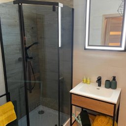 Bartosz Górski - Nowoczesna łazienka po remoncie: kabina prysznicowa typu walk-in z czarnymi profilami, umywalka z szafką i podświetlane lustro. Wykończenie w beżach i czerni.