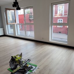 Bartosz Górski - Jasne panele podłogowe w pokoju z oknami, przez które widać budynek. Na podłodze piła do paneli na kartonie. Nowoczesna lampa sufitowa.