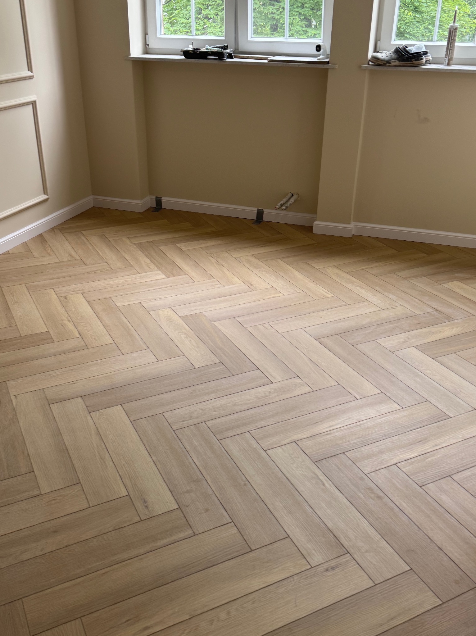 Jasna, drewniana podłoga ułożona w jodełkę w pokoju z beżowymi ścianami i białymi listwami przypodłogowymi. Na parapecie okna widoczne akcesoria malarskie.