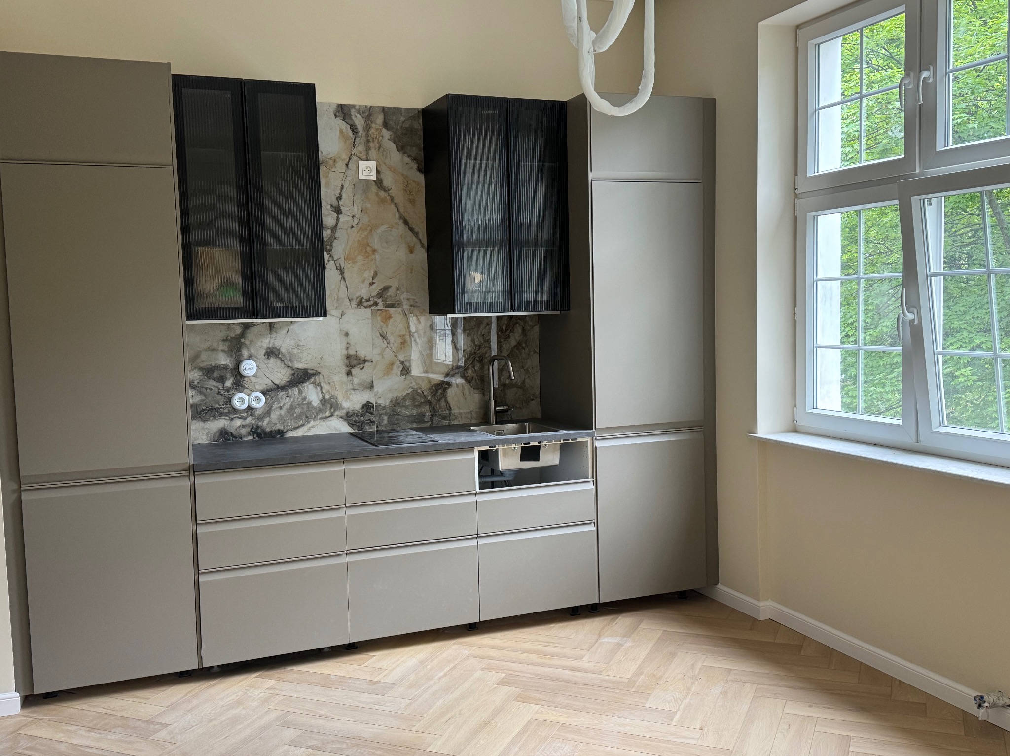 Ukończona kuchnia w odcieniach szarości z marmurowym backsplashiem i czarnymi szafkami wiszącymi. Widok na okno z zielenią za nim i parkiet w jodełkę.