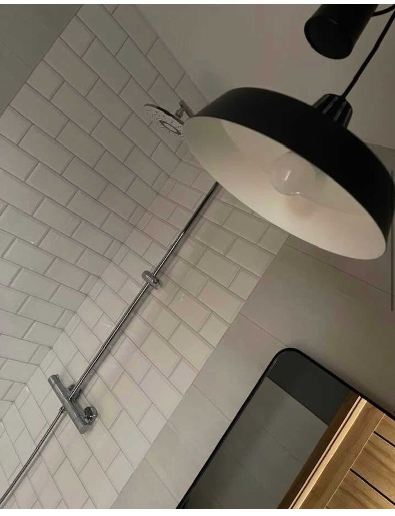 Nowoczesna łazienka z białymi płytkami, chromowaną armaturą prysznicową i czarną lampą wiszącą. Fragment lustra z drewnianą ramą w rogu kadru.