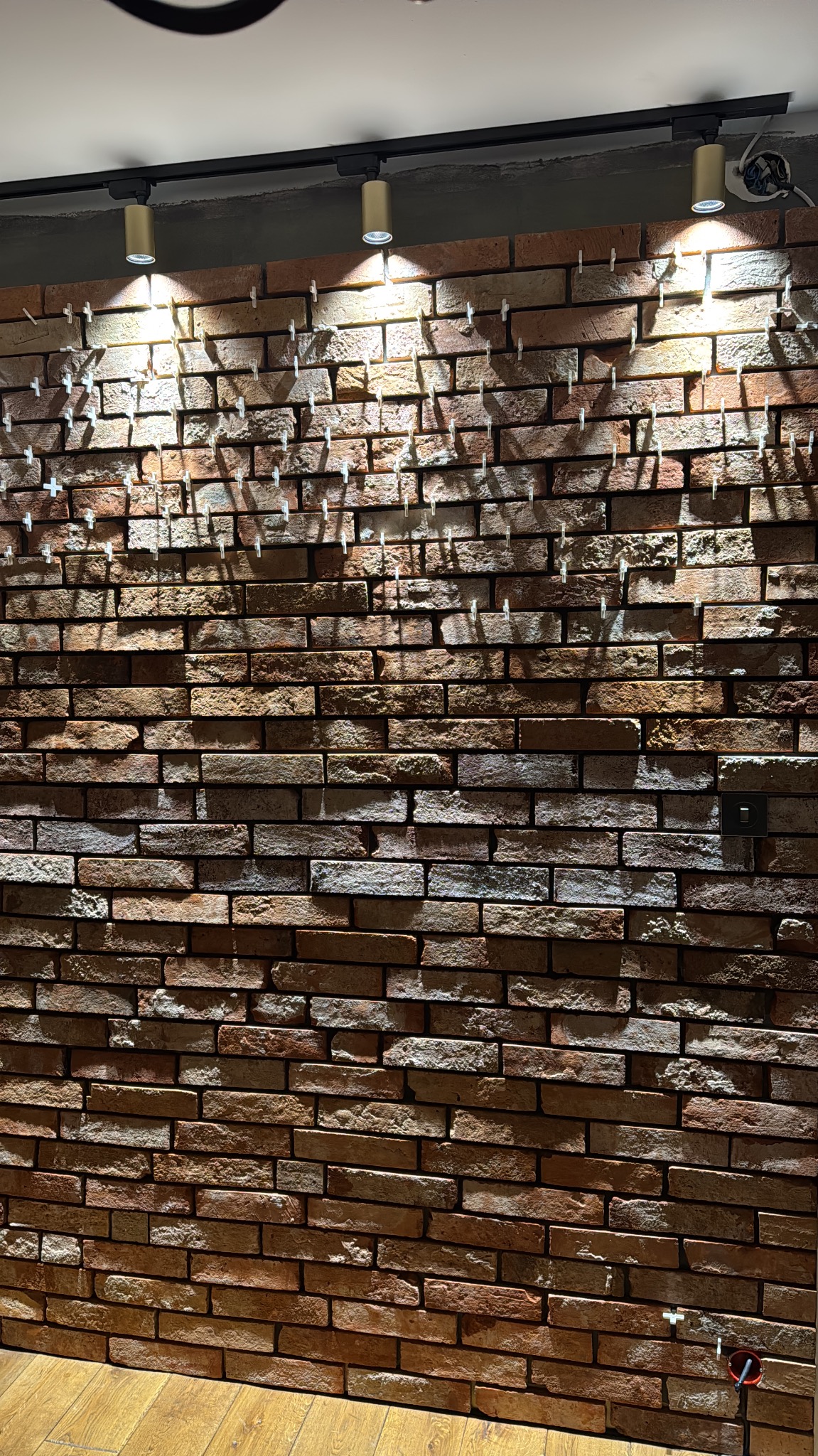 Ściana z cegły dekoracyjnej, oświetlona reflektorami szynowymi. Widoczne krzyżyki dystansowe między cegłami. Podłoga drewniana, fragment instalacji elektrycznej.