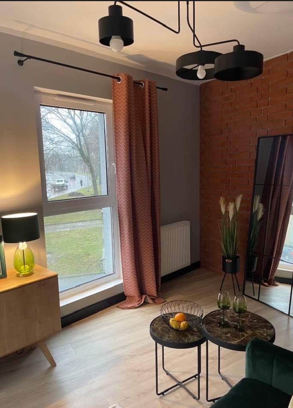Stylowe wnętrze: Okno z wzorzystymi zasłonami, nowoczesna lampa sufitowa, ściana z cegły i dekoracyjne stoliki kawowe. Elementy aranżacji wnętrz.