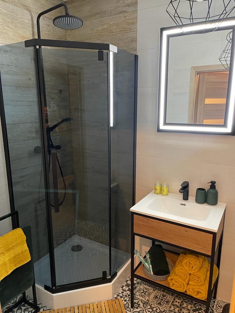 Nowoczesna łazienka po remoncie: kabina prysznicowa typu walk-in z czarnymi profilami, umywalka z szafką i podświetlane lustro. Wykończenie w beżach i czerni.