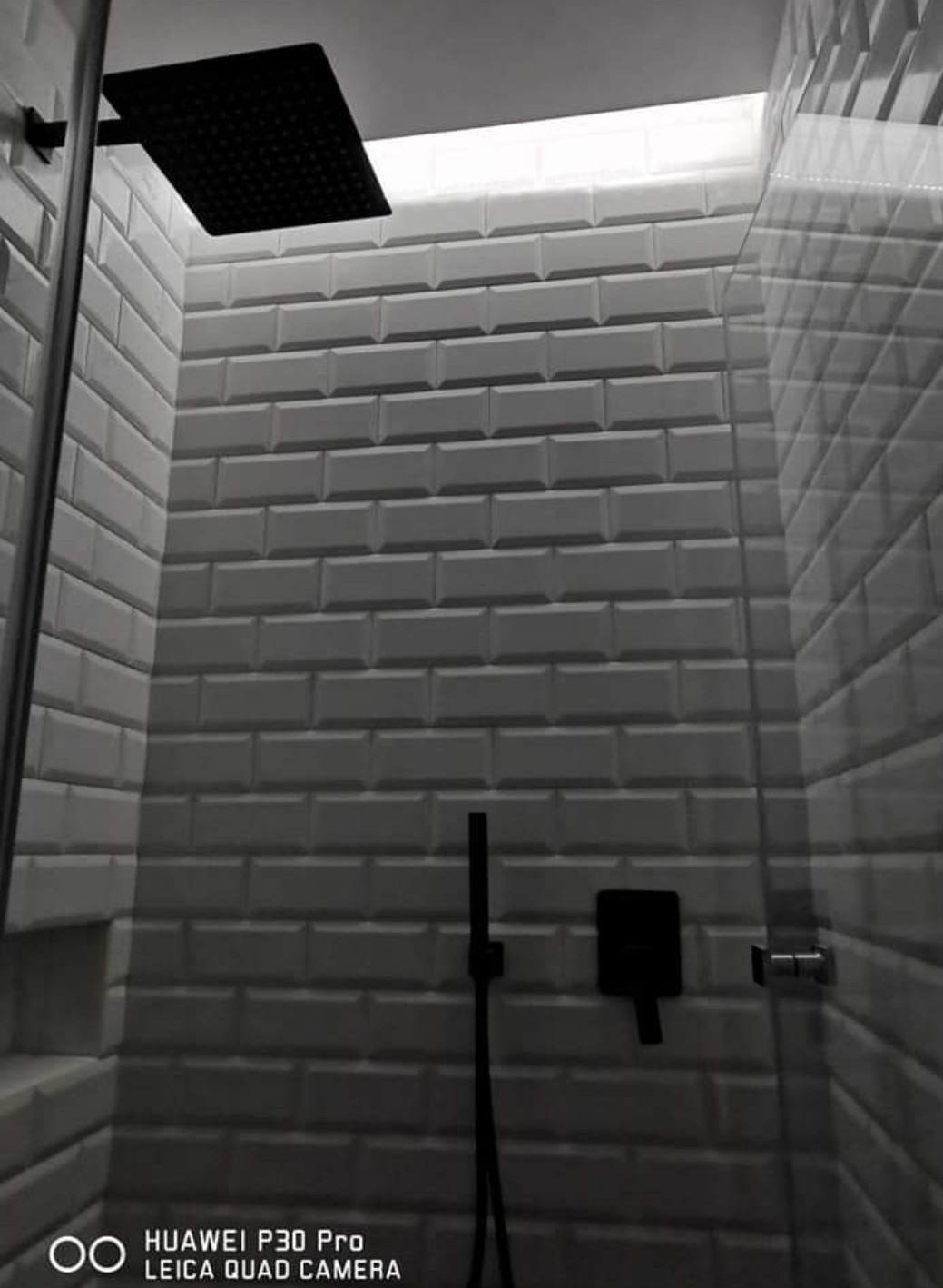 Nowoczesna kabina prysznicowa z czarną armaturą i białymi, wypukłymi płytkami w stylu cegiełek. Szklana ściana kabiny i oświetlenie LED pod sufitem.