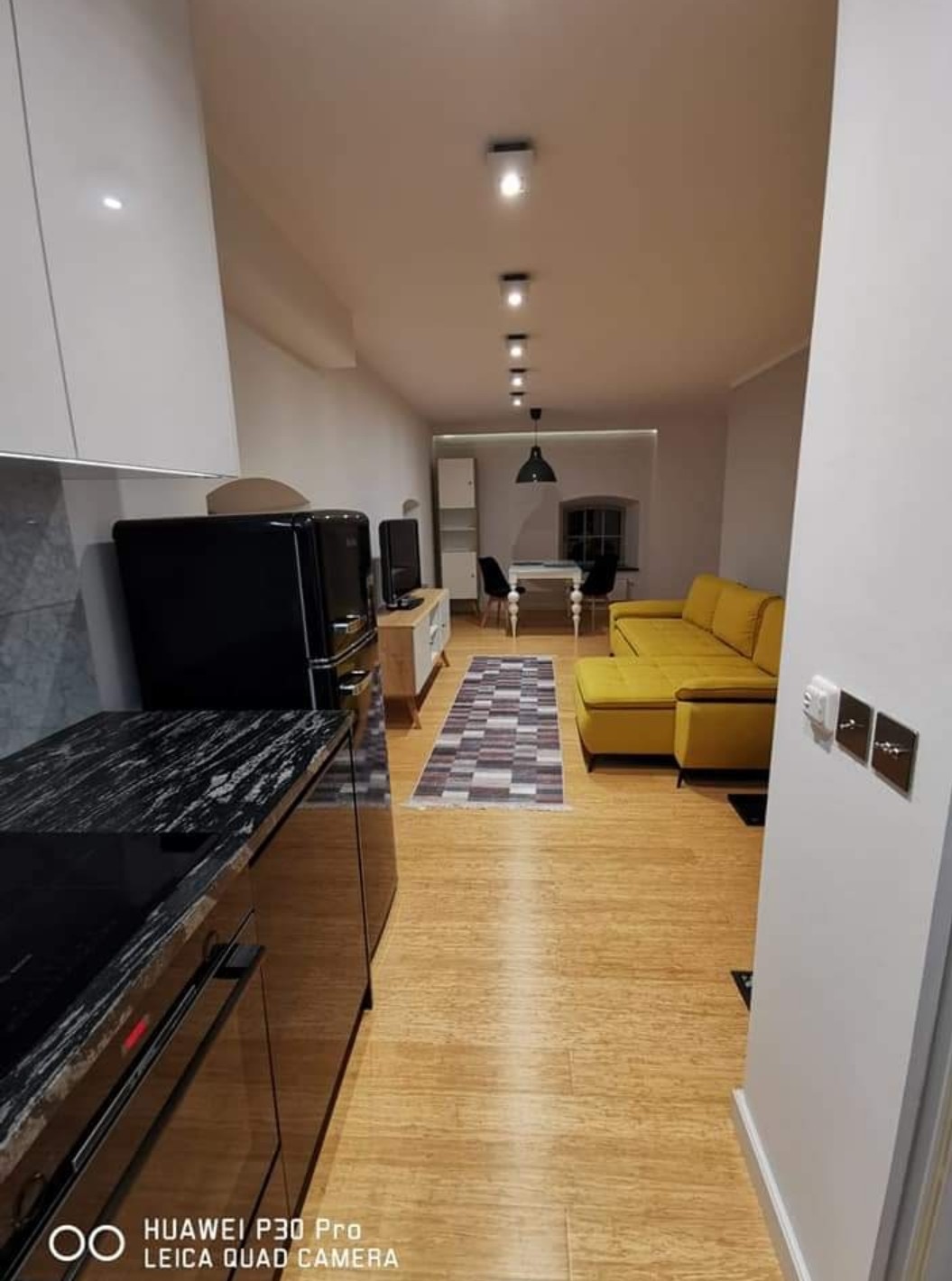 Nowoczesne wnętrze mieszkania po remoncie: kuchnia z czarnym blatem, żółta sofa, drewniana podłoga i oświetlenie punktowe. Widok z perspektywy wejścia.