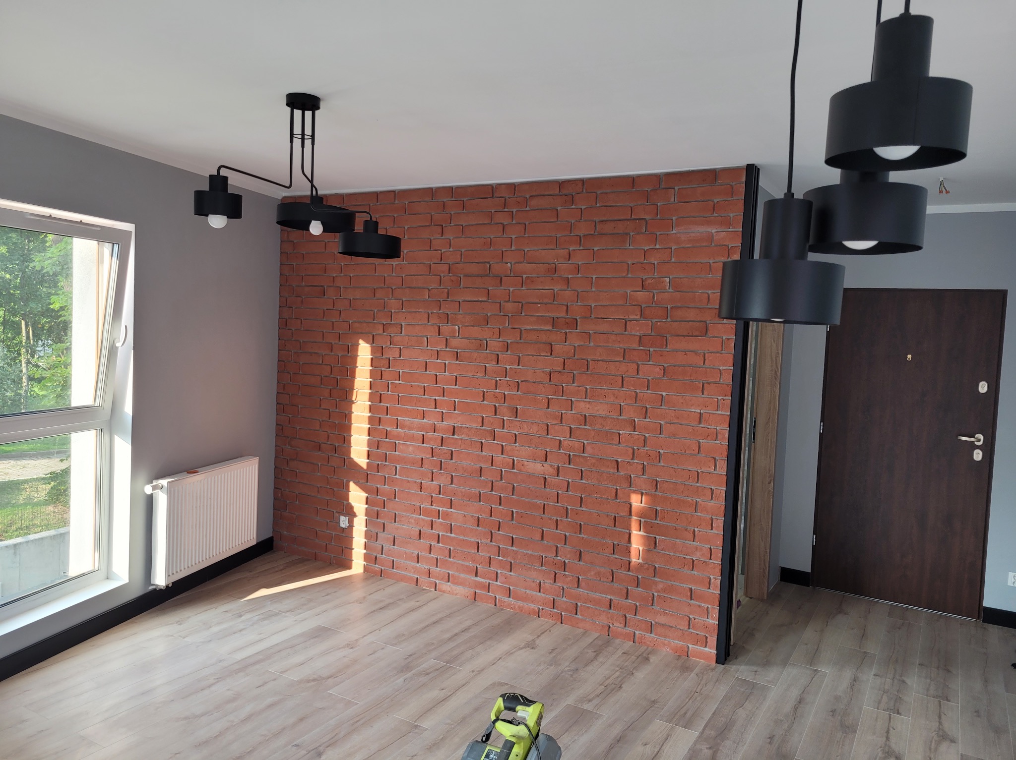 Loftowe wnętrze: ceglana ściana z czarną ramą, drewniana podłoga, nowoczesne lampy. Słoneczne światło podkreśla fakturę cegły. Widoczny fragment narzędzia.