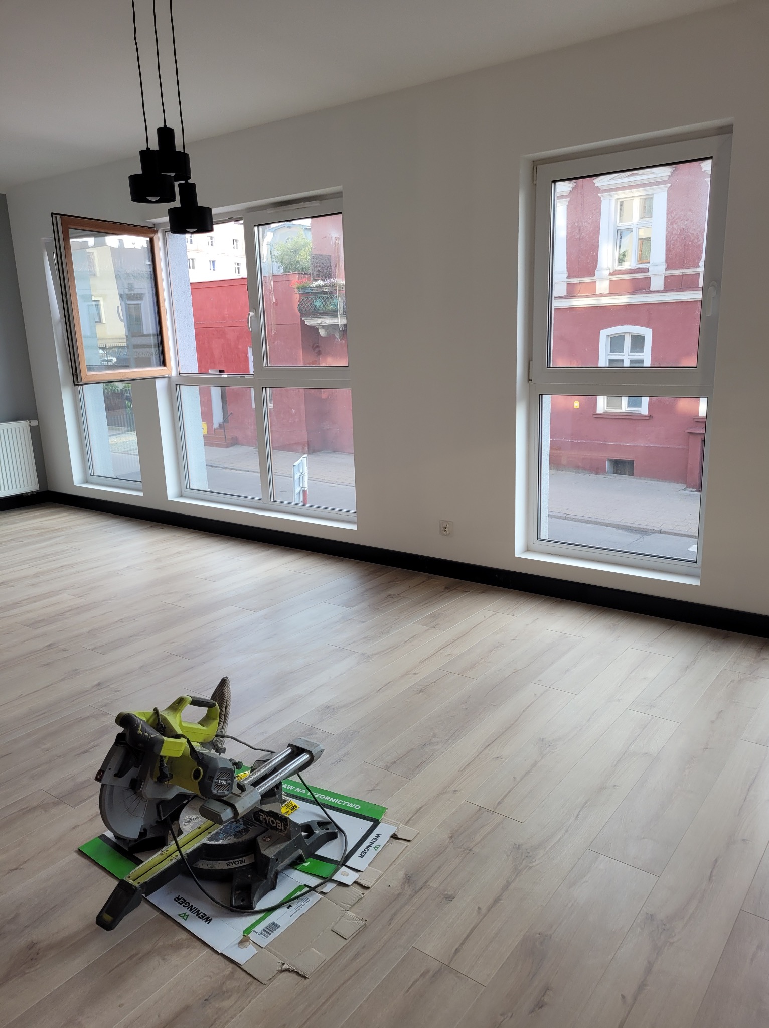 Jasne panele podłogowe w pokoju z oknami, przez które widać budynek. Na podłodze piła do paneli na kartonie. Nowoczesna lampa sufitowa.