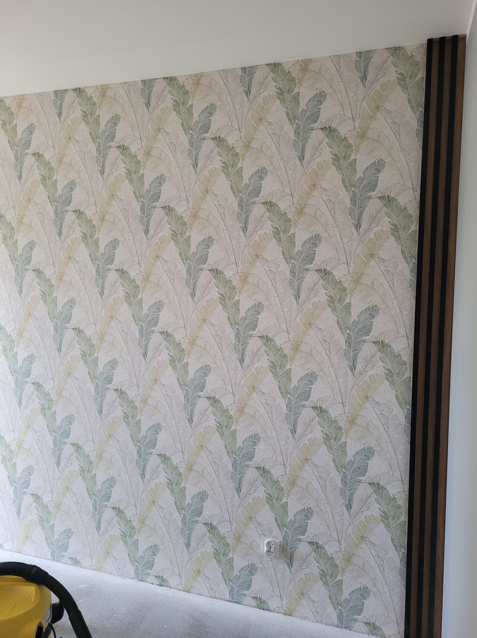 Ściana po tapetowaniu w Starym Dzierzgoniu z wzorem liści i dekoracyjnymi panelami ściennymi. Widoczny fragment odkurzacza.
