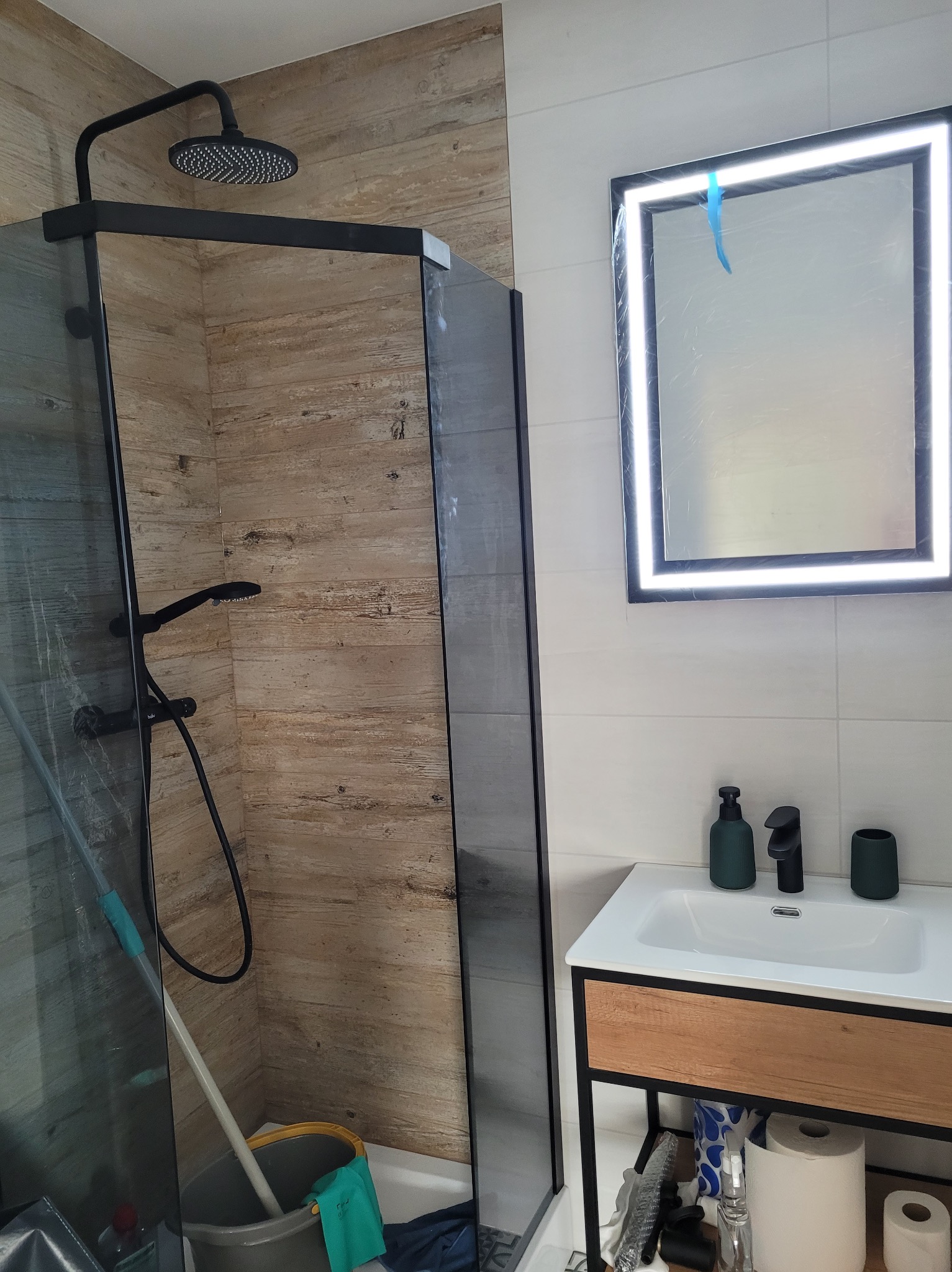 Nowoczesna łazienka po remoncie: kabina prysznicowa typu walk-in z czarną armaturą i podświetlane lustro nad umywalką z szafką. Widoczne akcesoria łazienkowe.