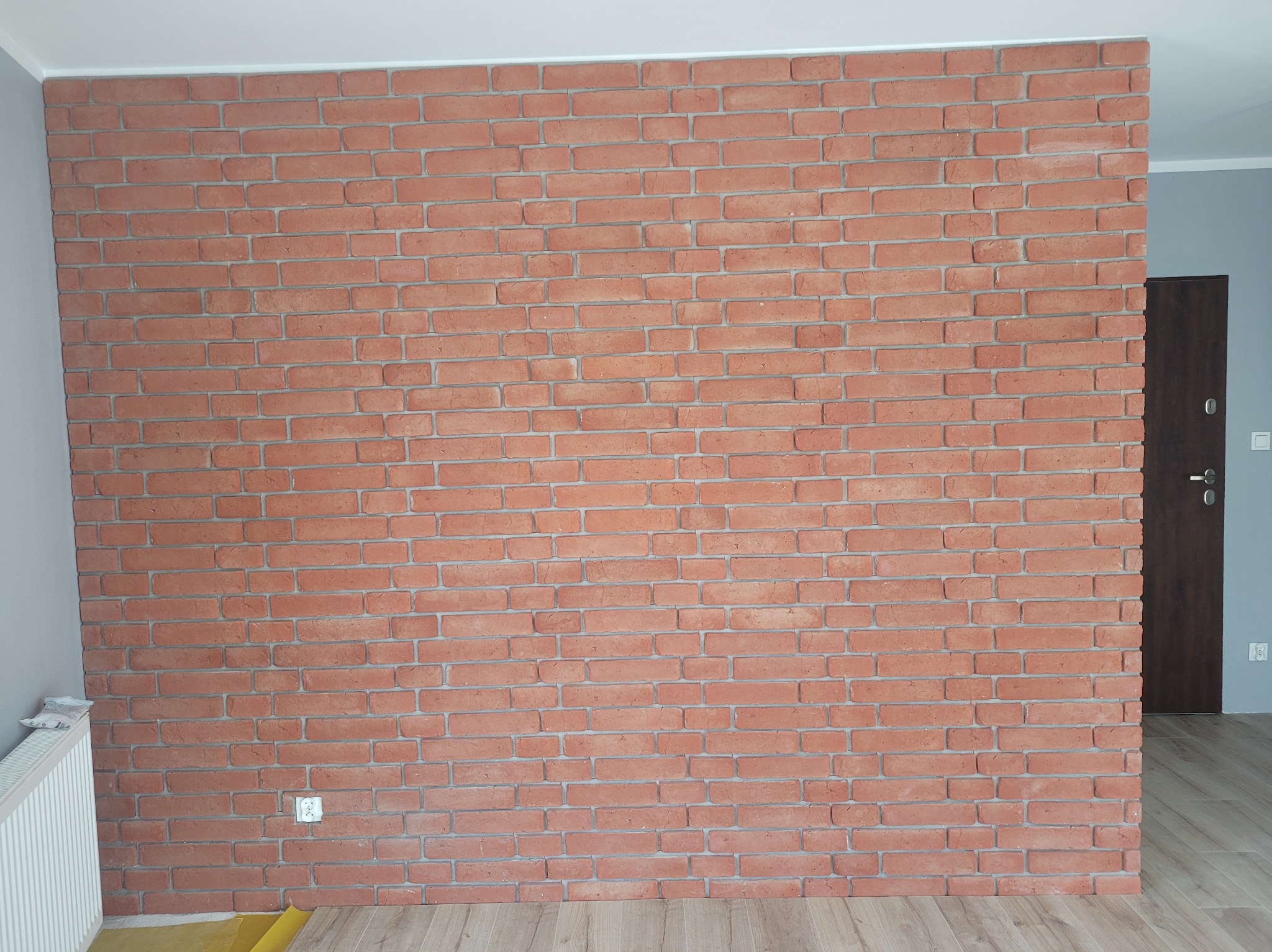 Dekoracyjna ściana z cegły w odcieniach czerwieni, wykończona szarą fugą. Widoczny fragment podłogi drewnopodobnej i gniazdko elektryczne.